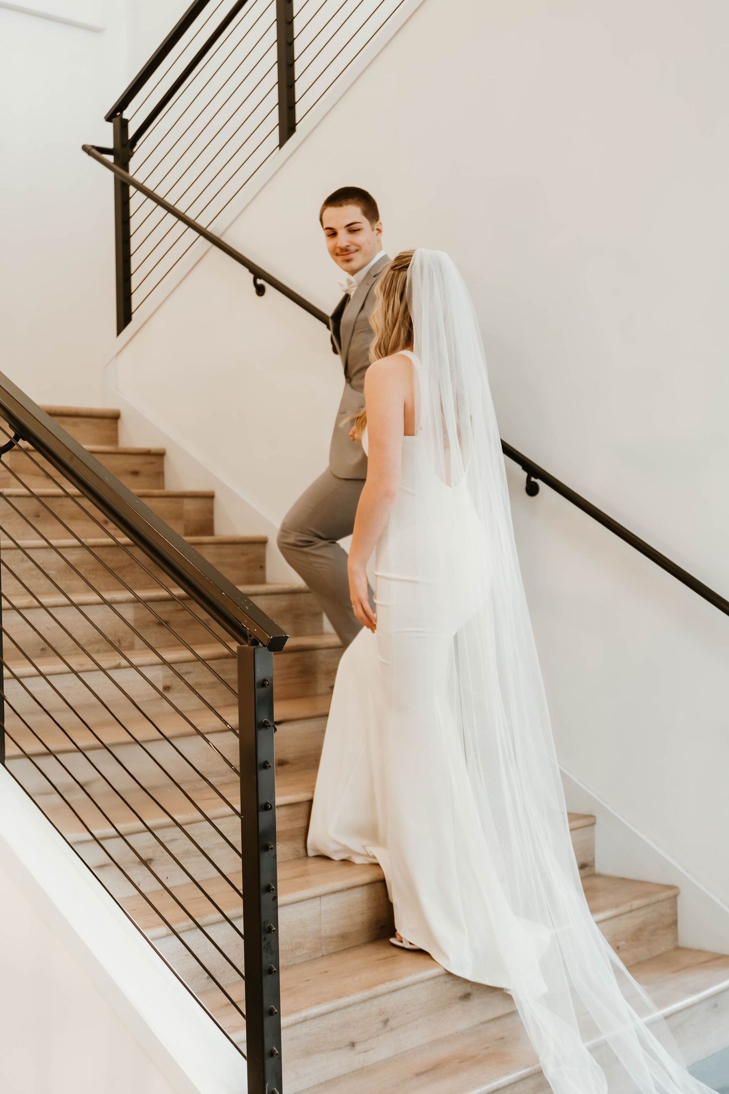 Bride in flowing white dress and veil ascending wooden staircase with metal railing.