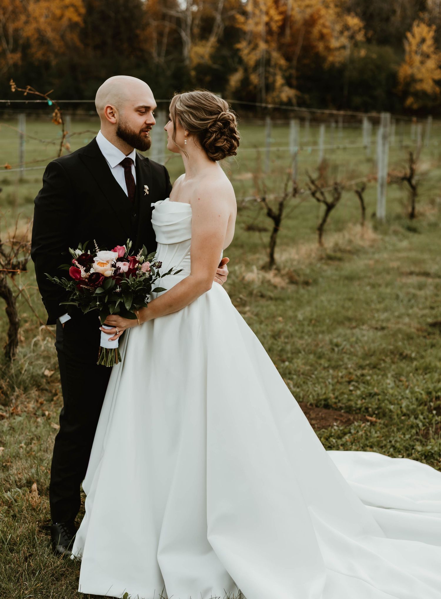 A bride in a flowing white gown stands in a rustic vineyard setting during fall.