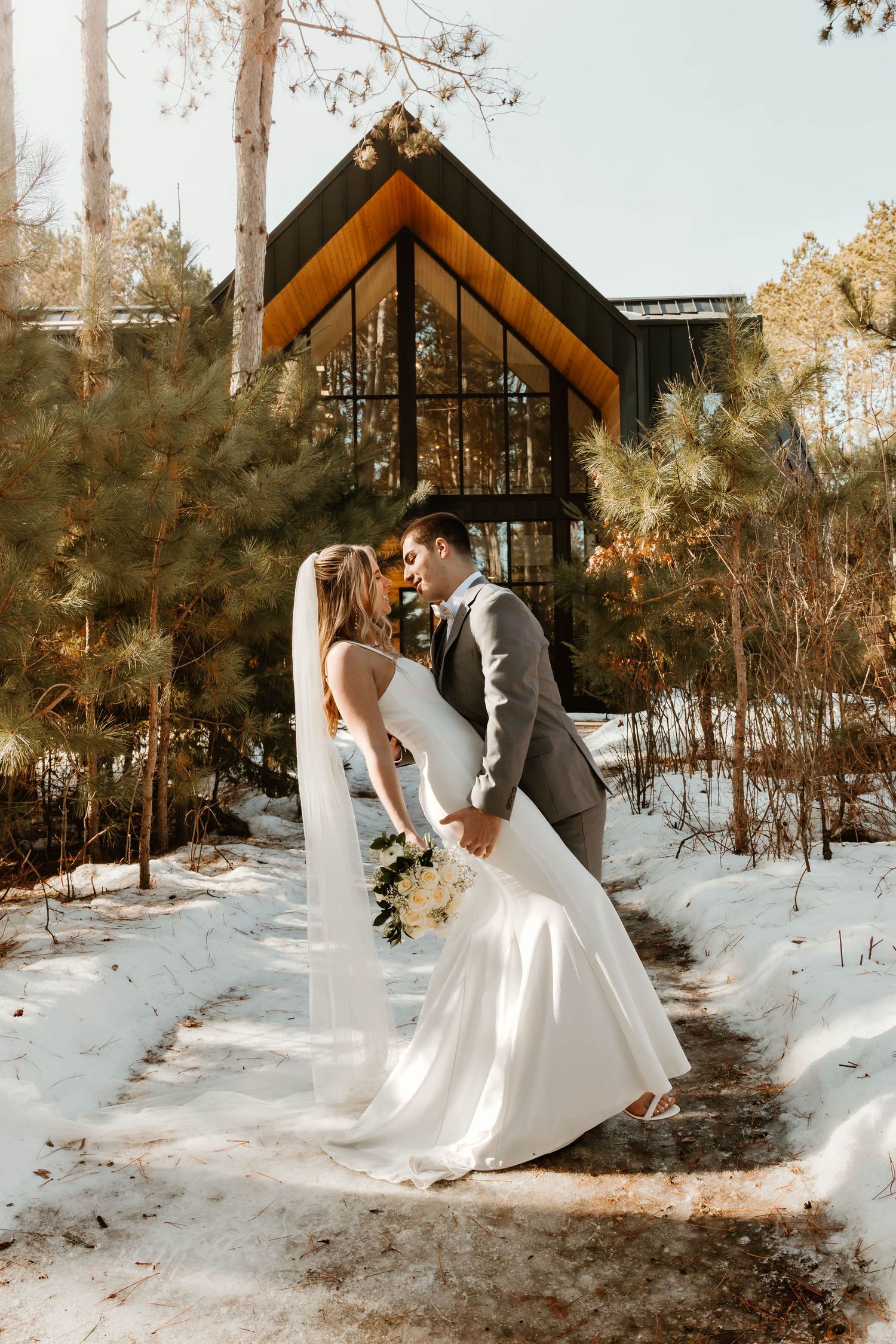 Romantic winter wedding photos in front of rustic wooden chapel with snow-covered grounds.
