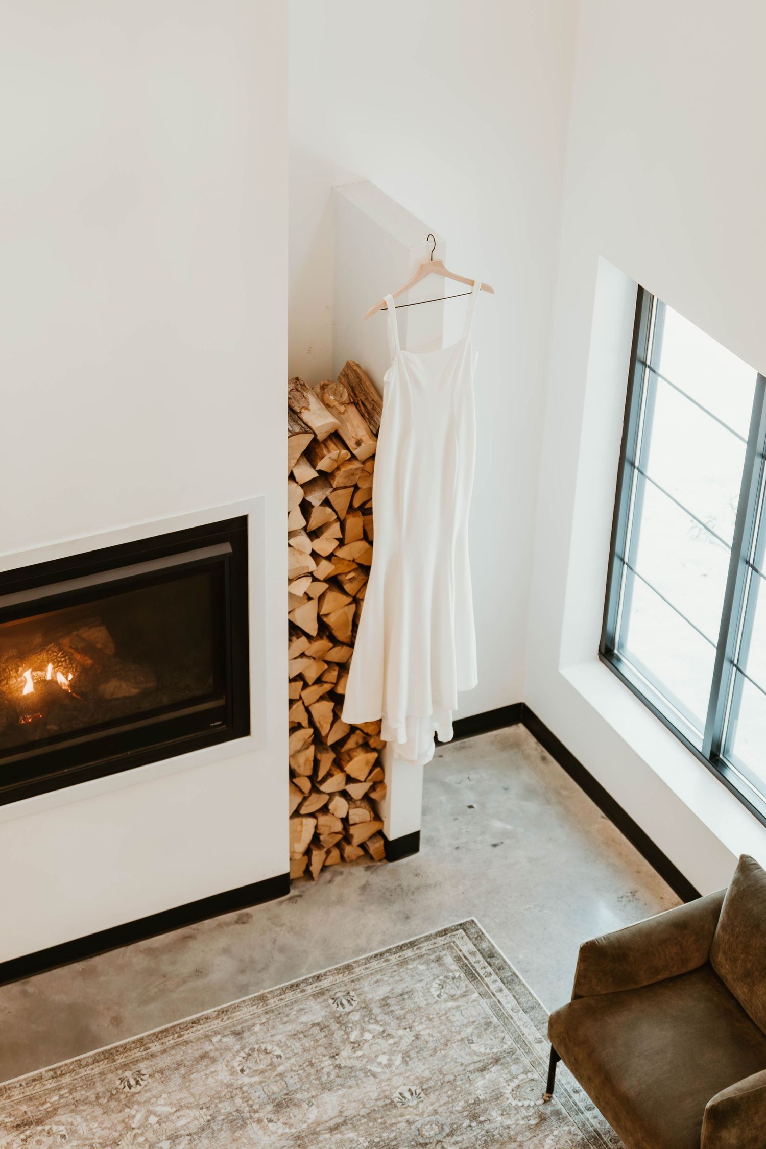 Wedding dress displayed near modern fireplace with stacked wood and large window.