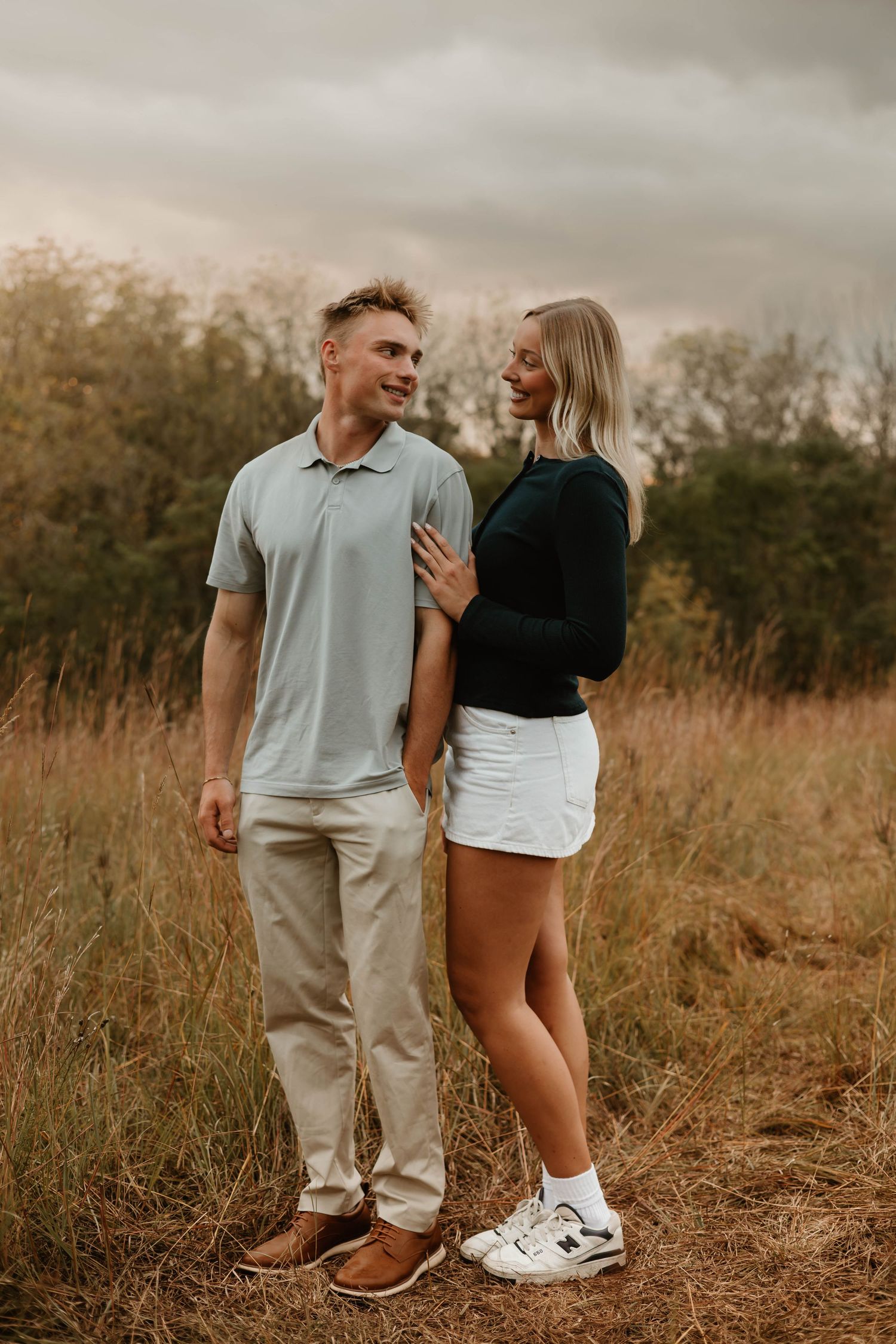 A couple stands close together in a golden field during sunset wearing casual attire and sneakers.
