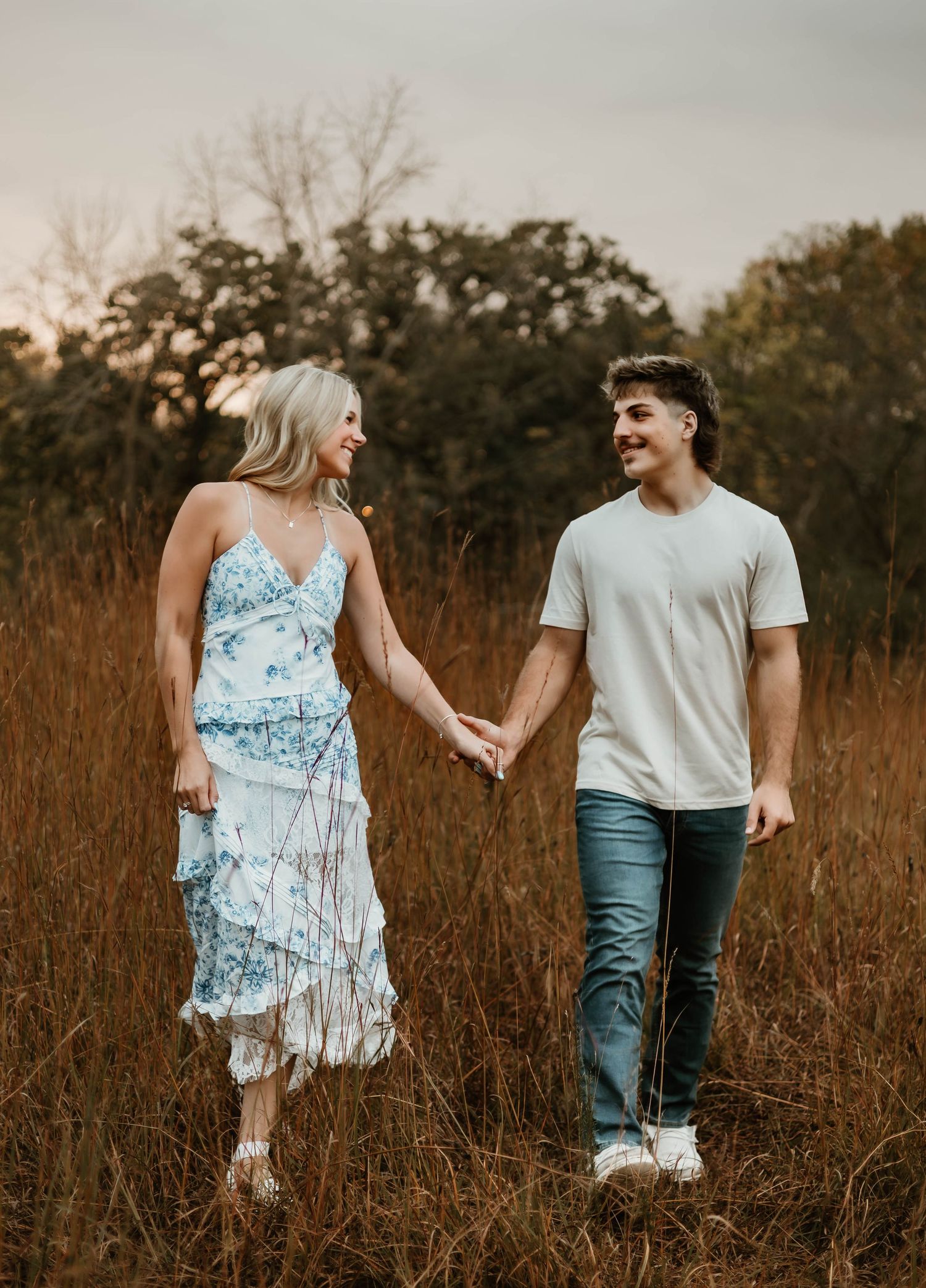 Couple holding hands while walking through a golden autumn field at sunset in casual attire.