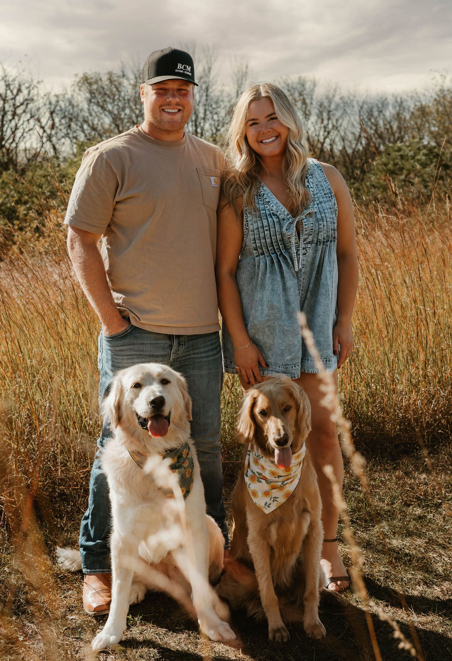 A couple poses with their two dogs in a golden field during sunset with tall grass in the background.