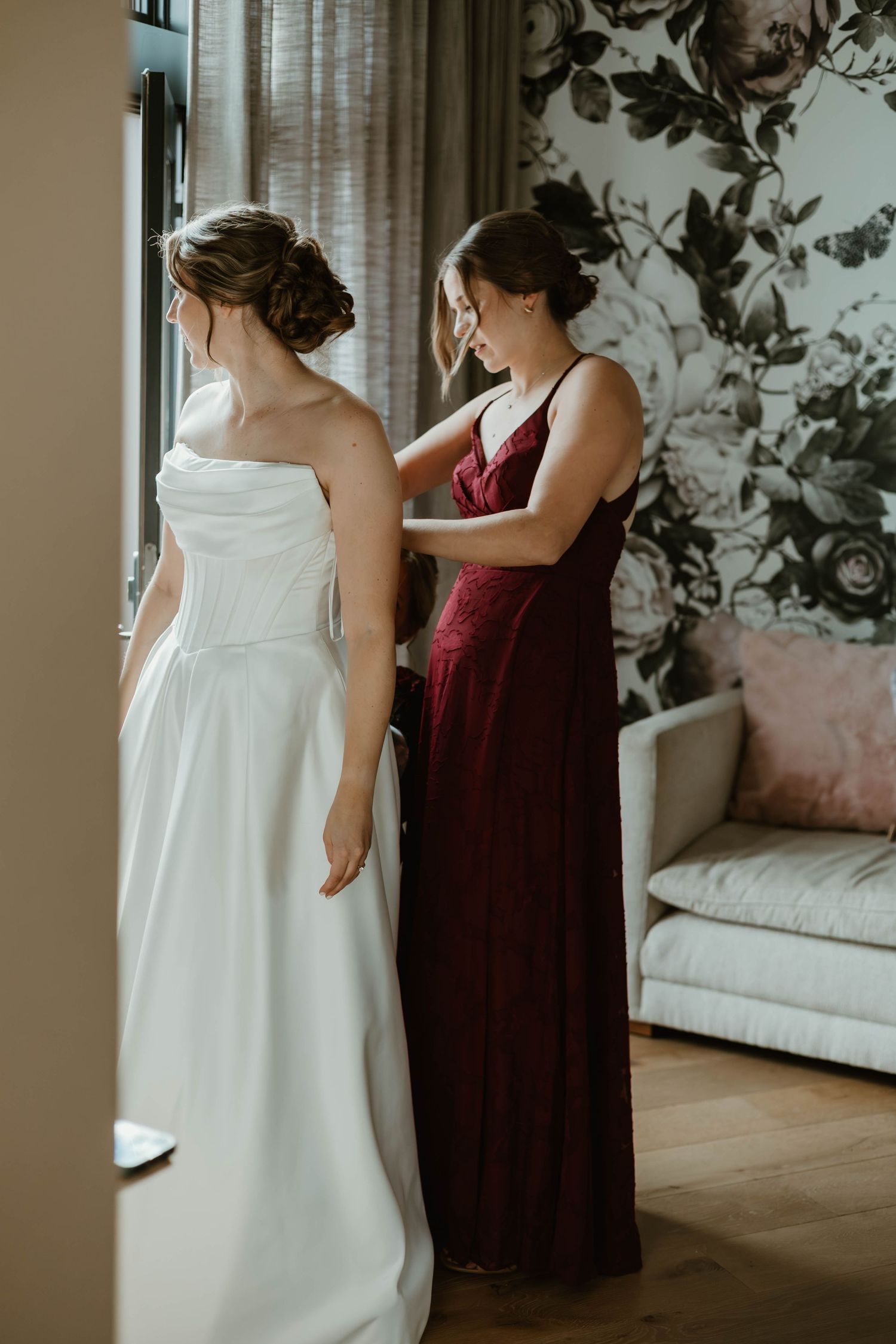 Two formal dresses shown side by side, one white and one burgundy, in an elegant room with floral wallpaper.