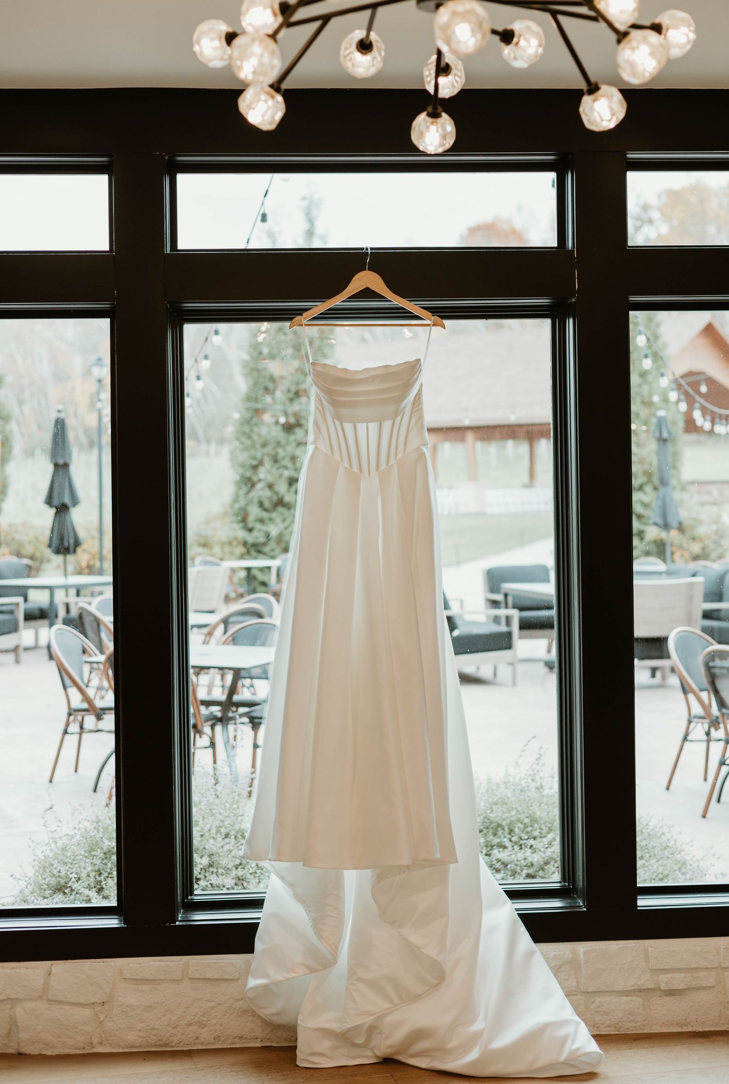Elegant white wedding dress hanging in front of large windows with natural light streaming in.