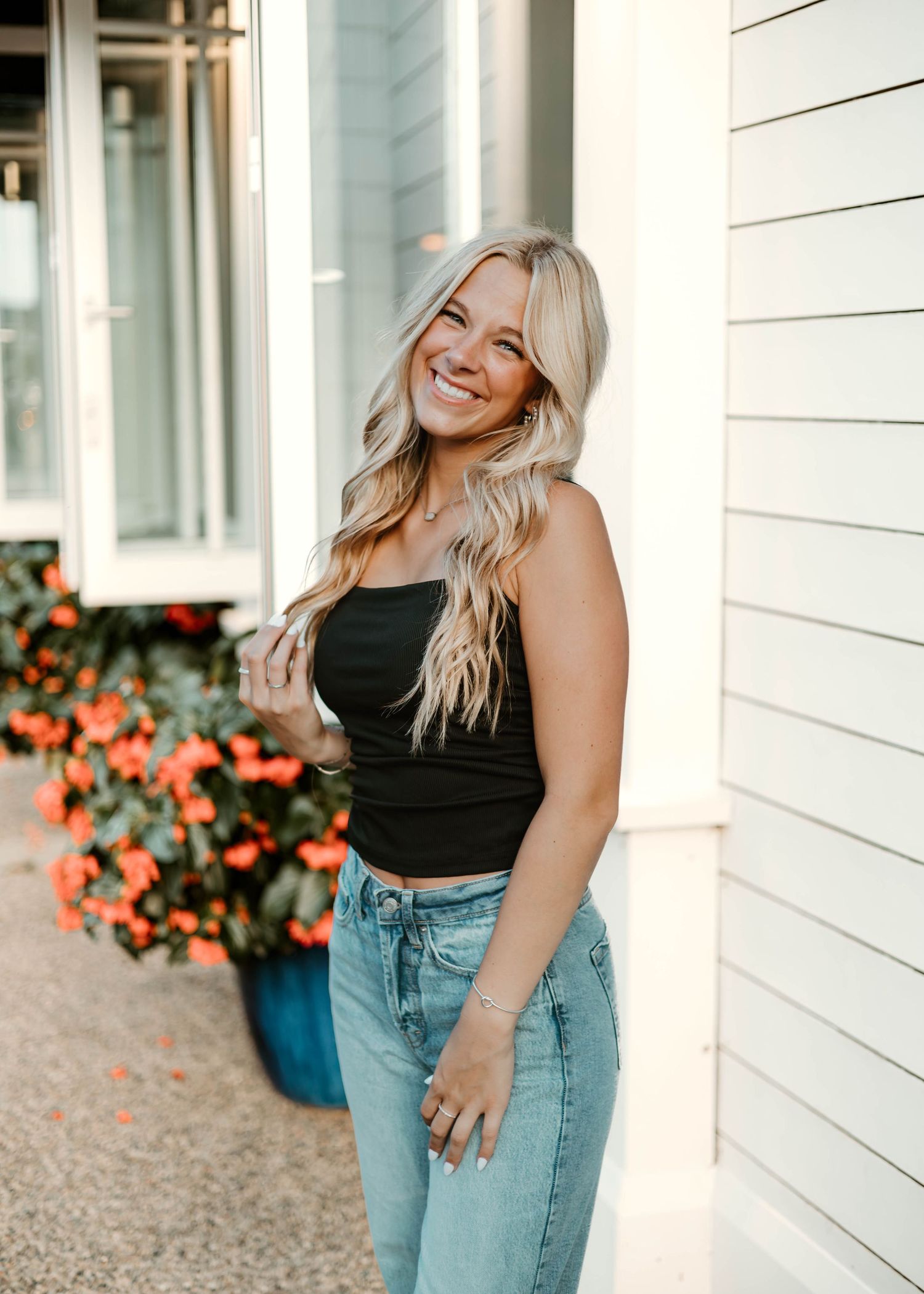 Smiling blonde woman in black crop top and light wash jeans posing outdoors near white siding and orange flowers.