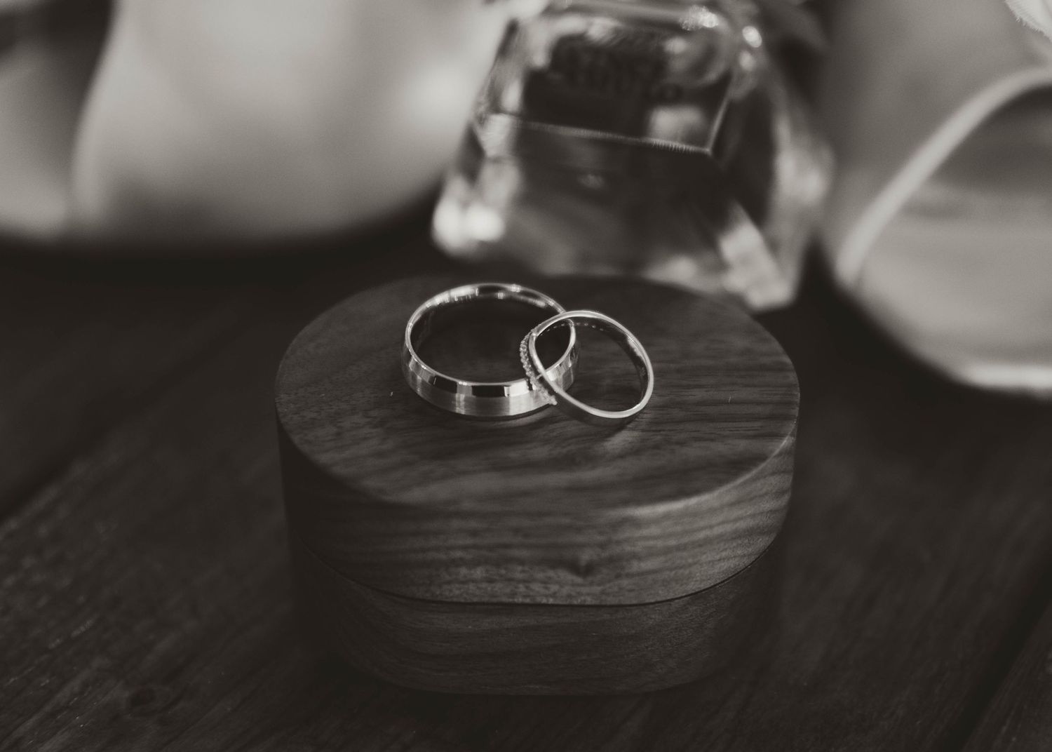 Two silver wedding rings rest on a dark wooden circular box in a black and white photograph.