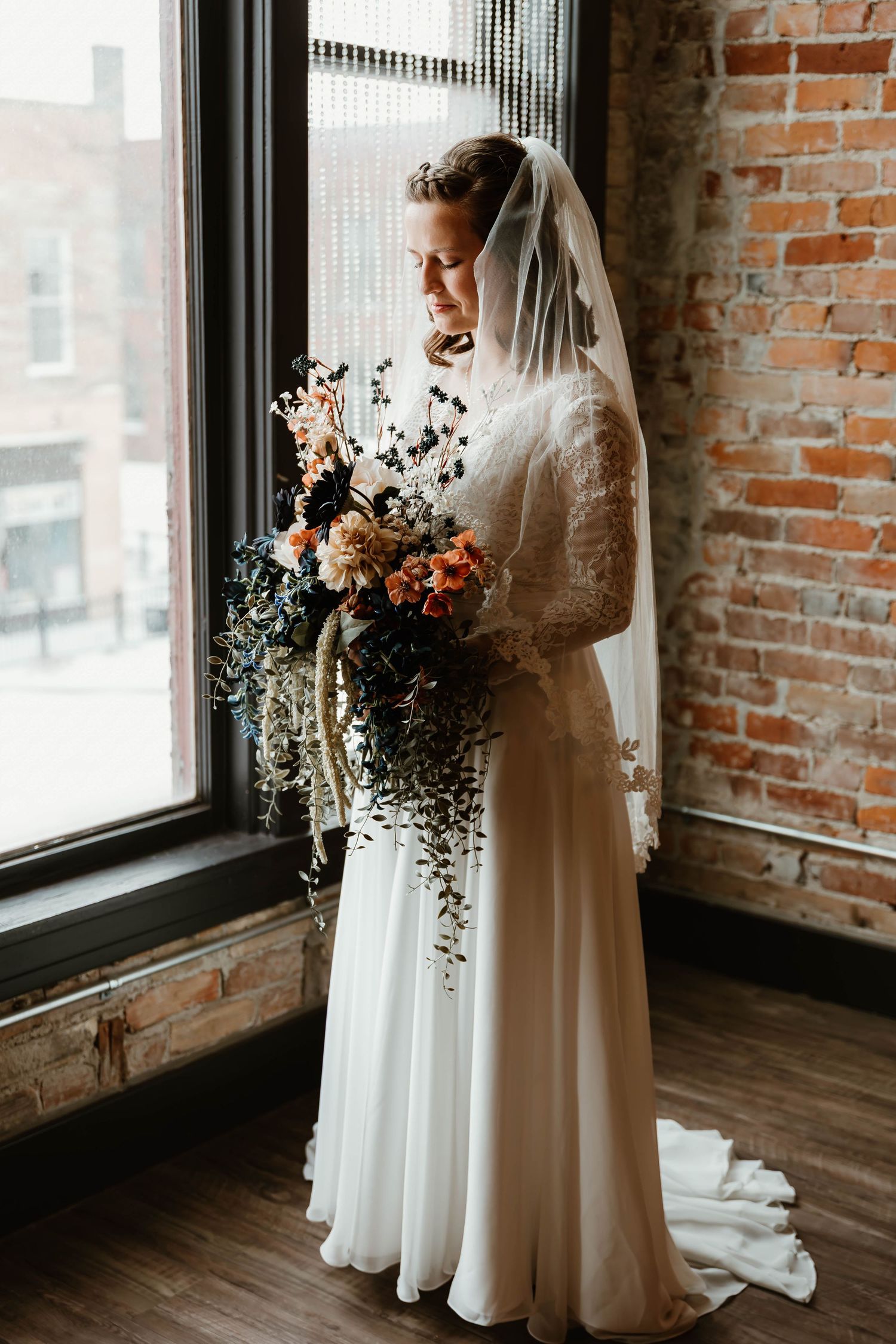 Elegant bridal portrait showing lace gown and cascading bouquet against brick wall and window.