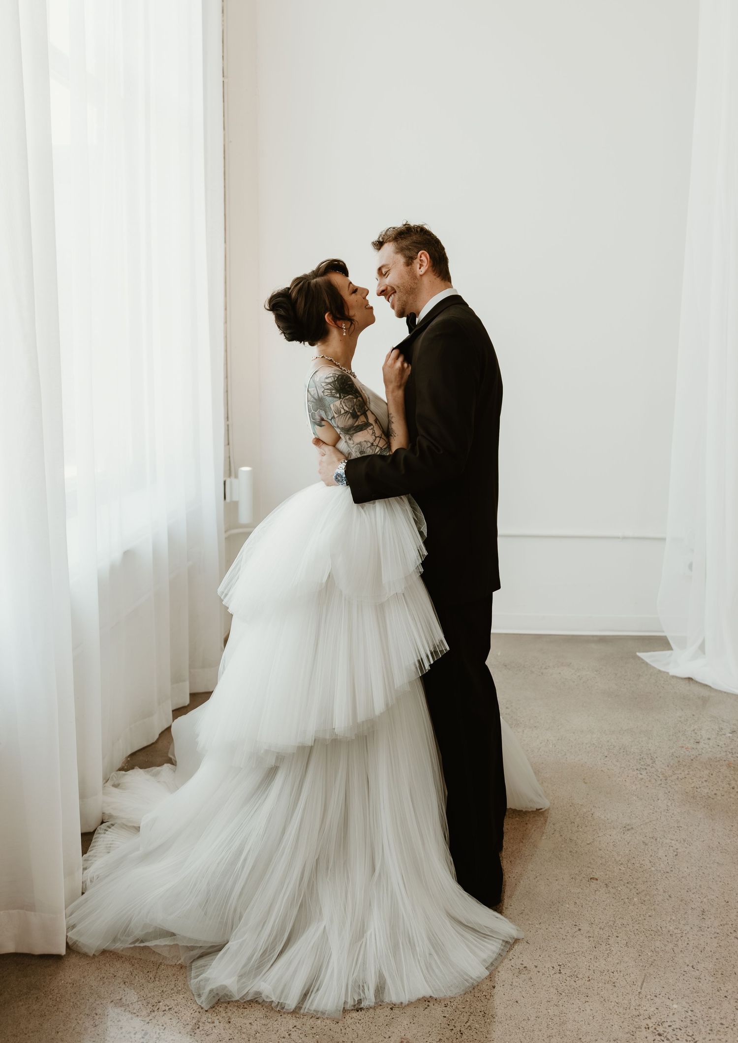 A bride in a tiered tulle gown and groom in a black tuxedo share a romantic moment by a white curtain.