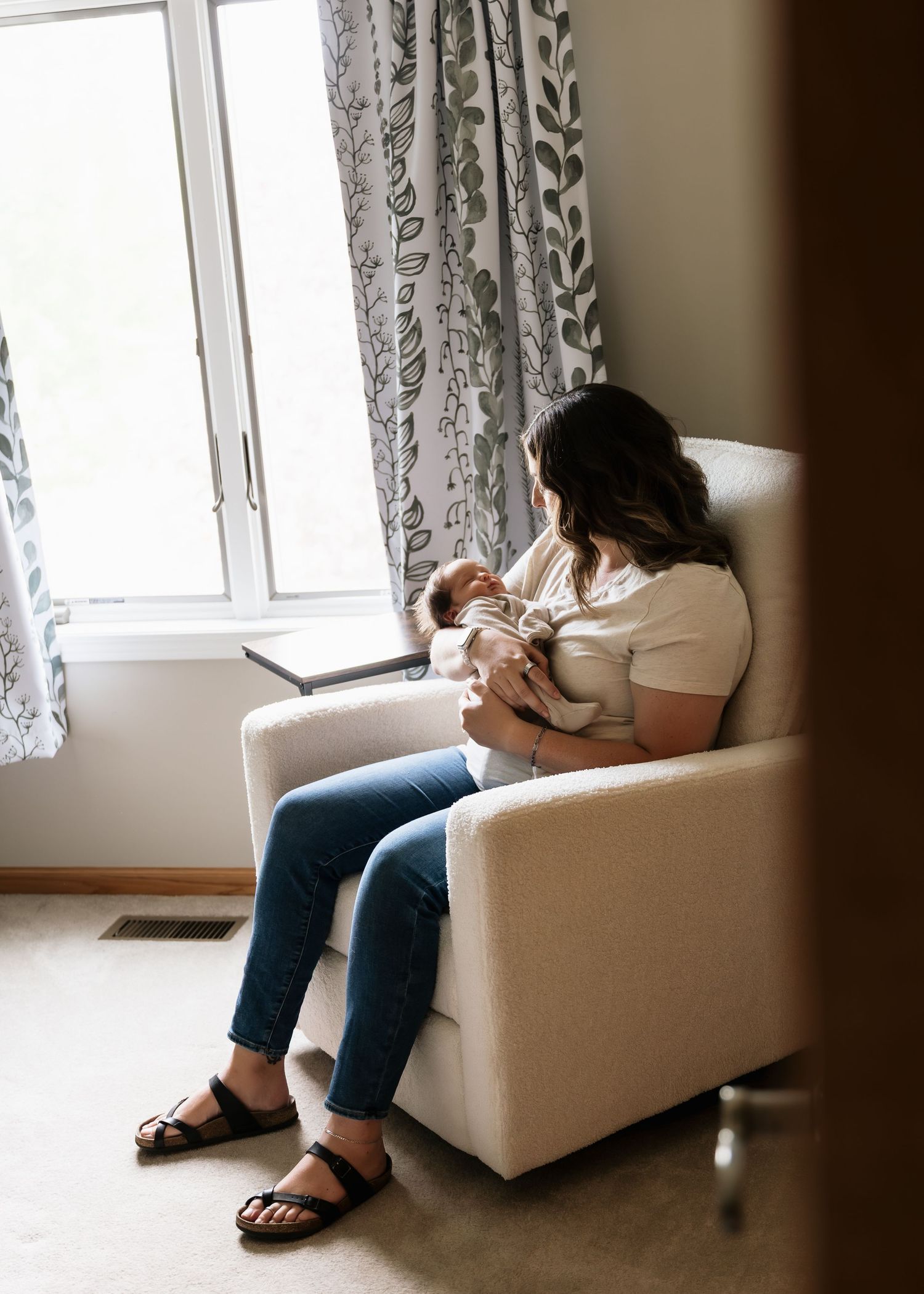 A mother sits in a white armchair by a window, cradling her newborn baby while breastfeeding in a cozy nursery.