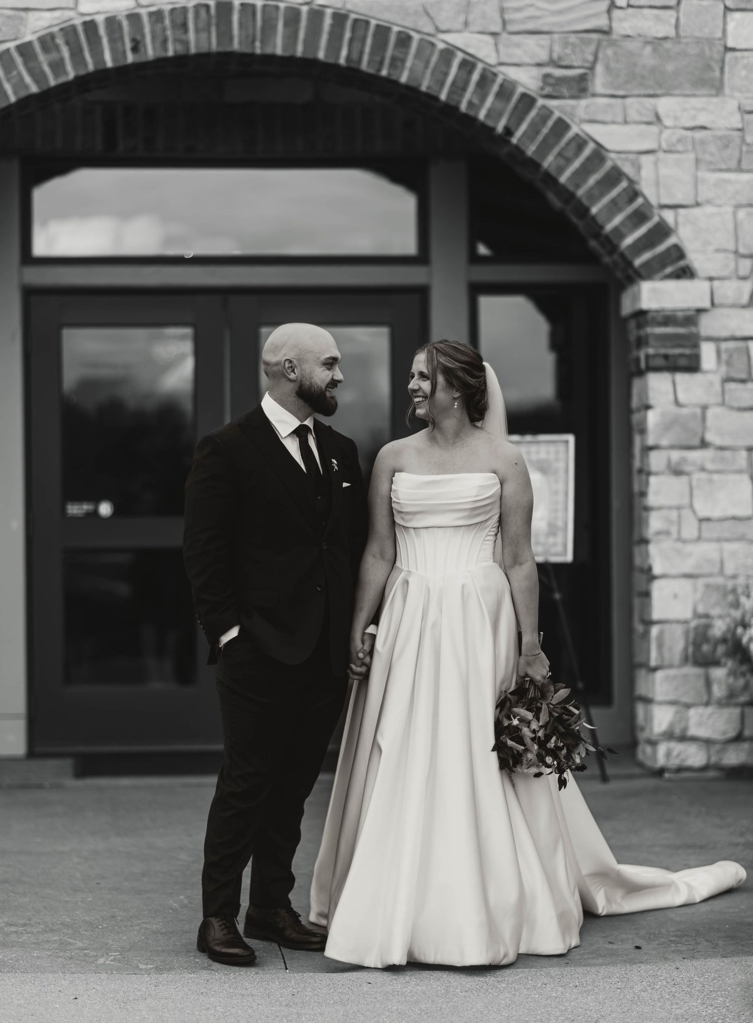 Elegant couple poses against stone archway building in classic black and white wedding portrait.