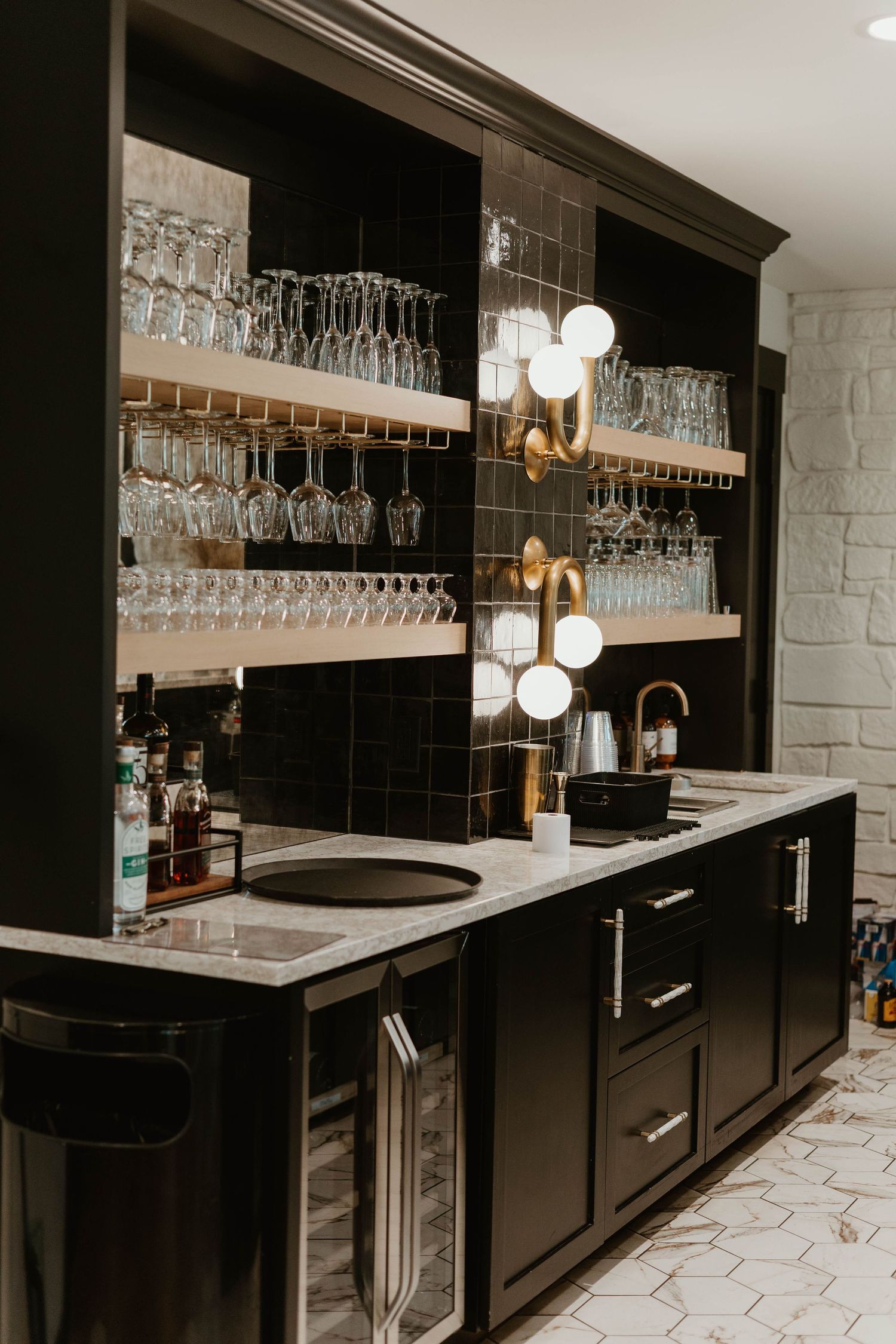 Modern home bar with black cabinets, marble countertops, and hanging glassware storage features elegant lighting fixtures.