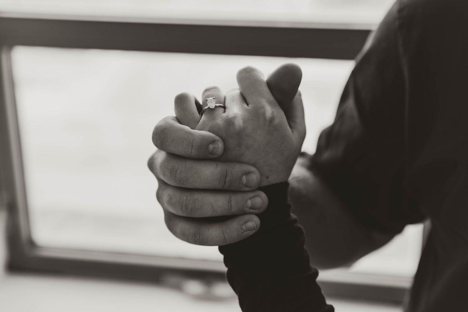 Black and white close-up photograph of hands clasped together in prayer or contemplation near a window.