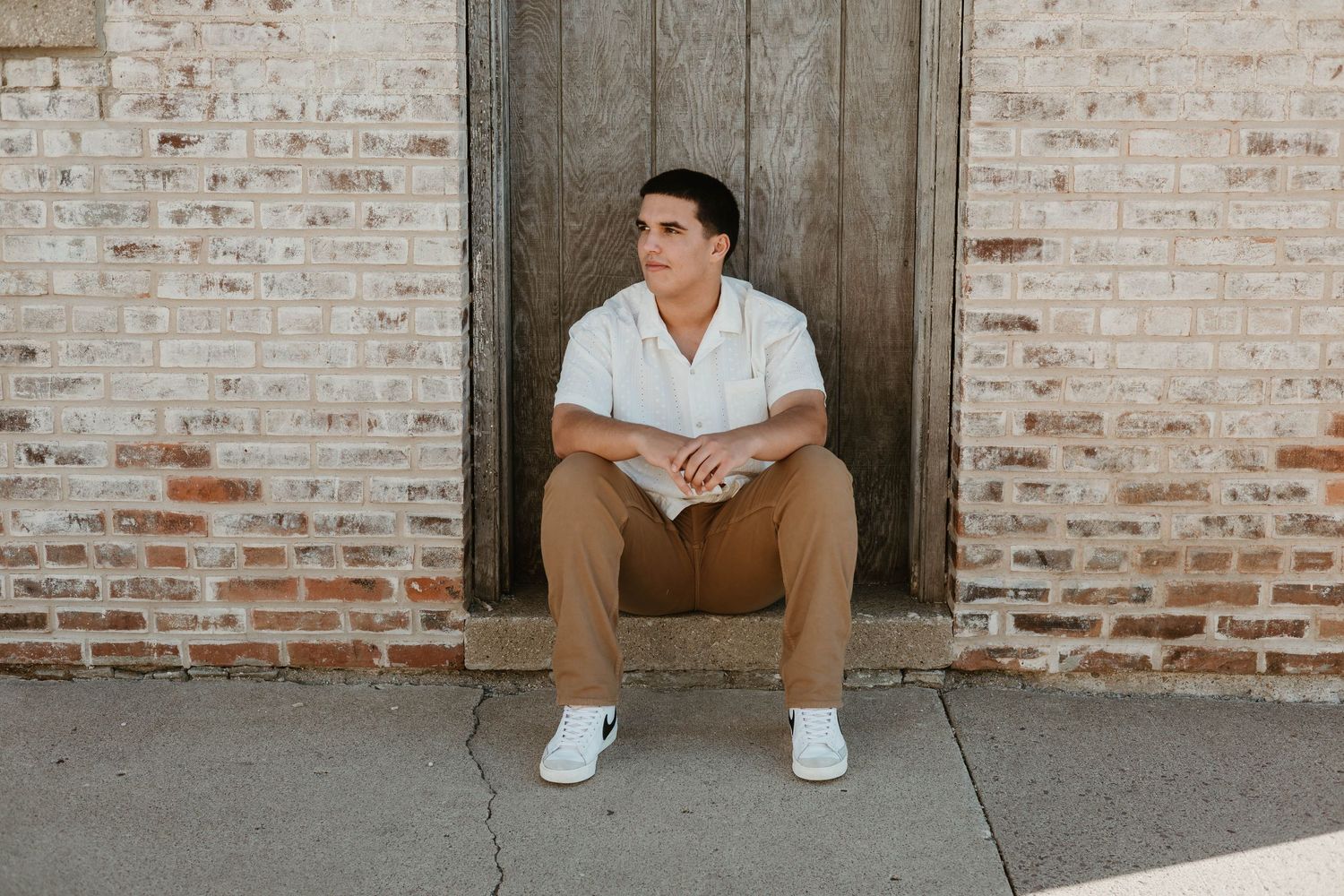 Young man in white shirt and tan pants sitting on steps against a brick wall and wooden door.
