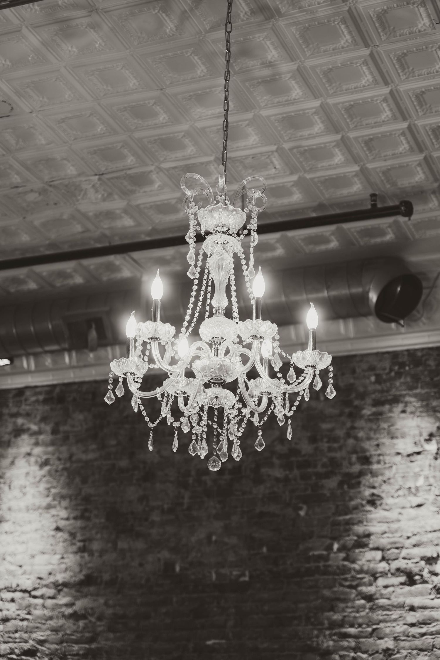 Crystal chandelier with lit candles hanging against exposed brick wall and tin ceiling.