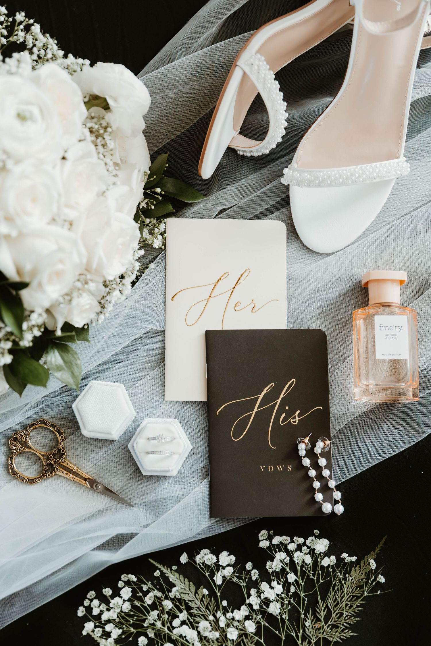 Wedding accessories including vows books, perfume, jewelry and white flowers on dark surface.