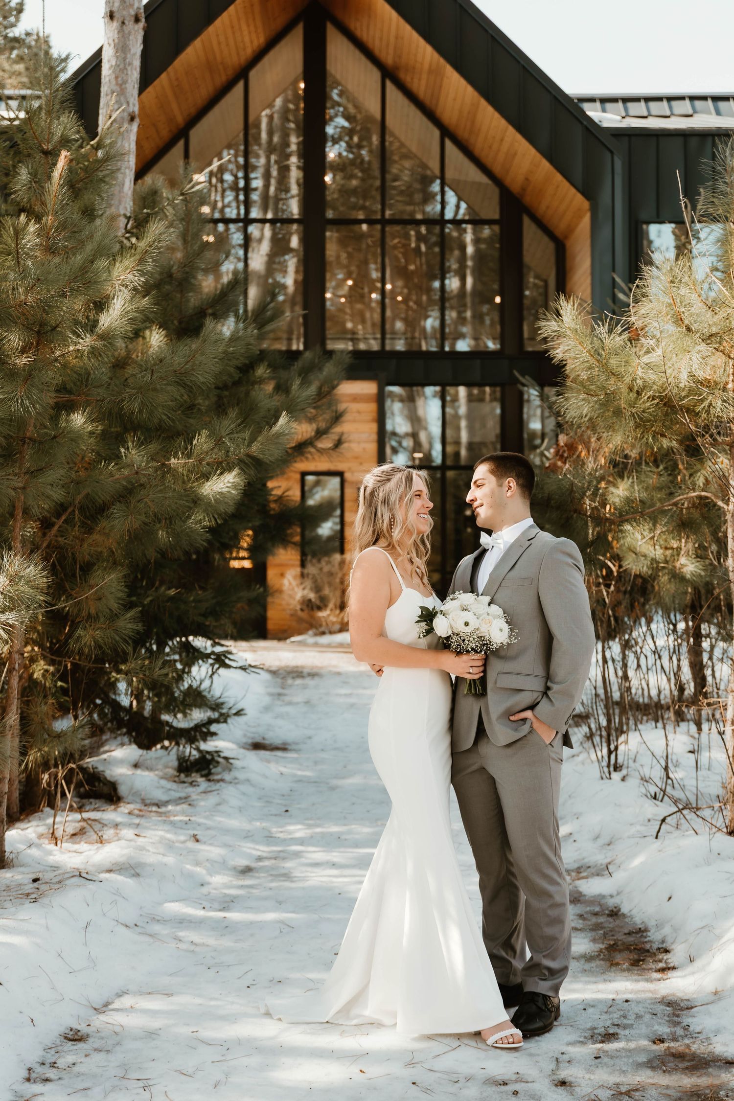 Winter wedding scene at rustic wooden chapel surrounded by snow and pine trees.