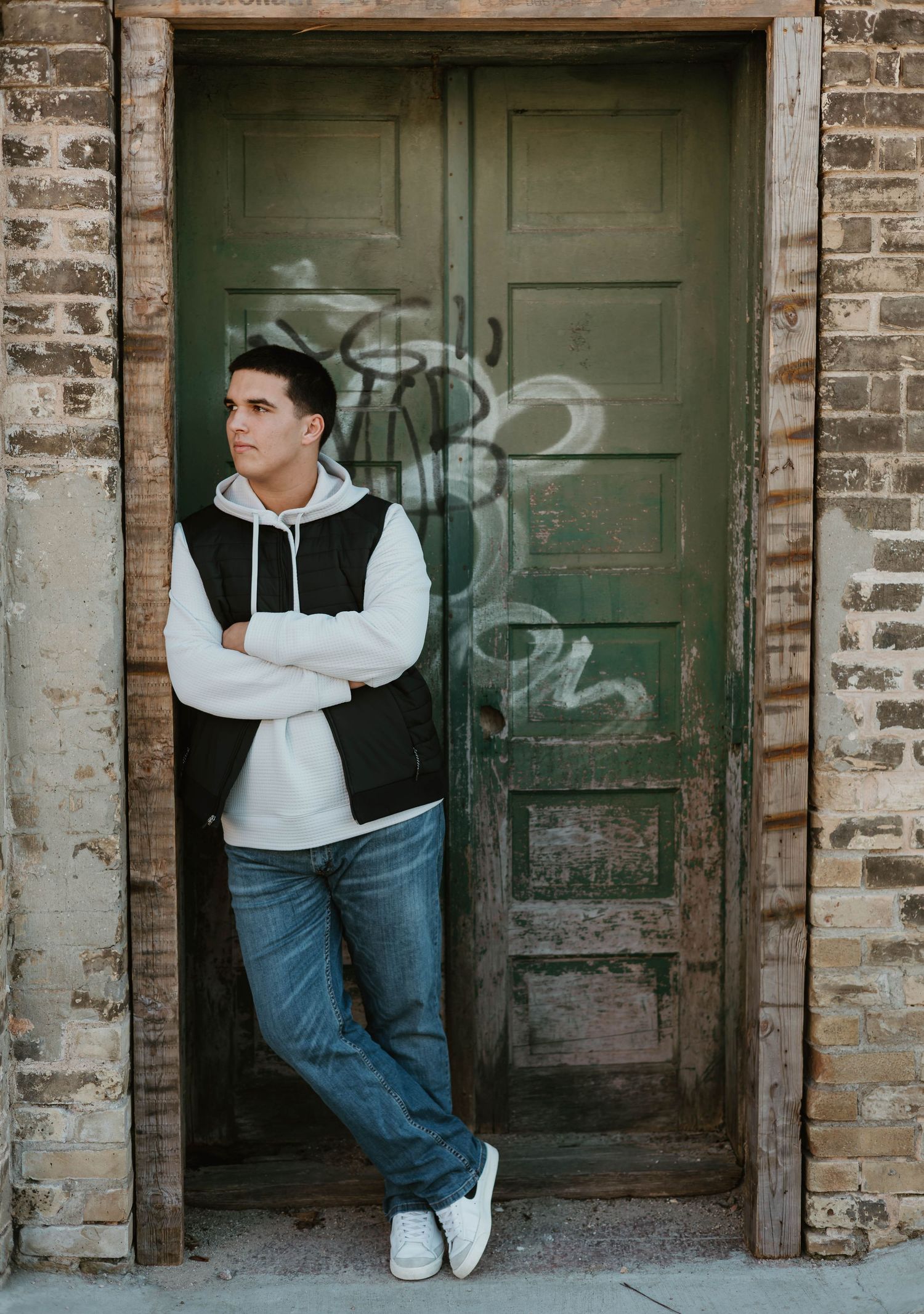 Young man in black vest and jeans leans against a green graffiti door in a brick building.