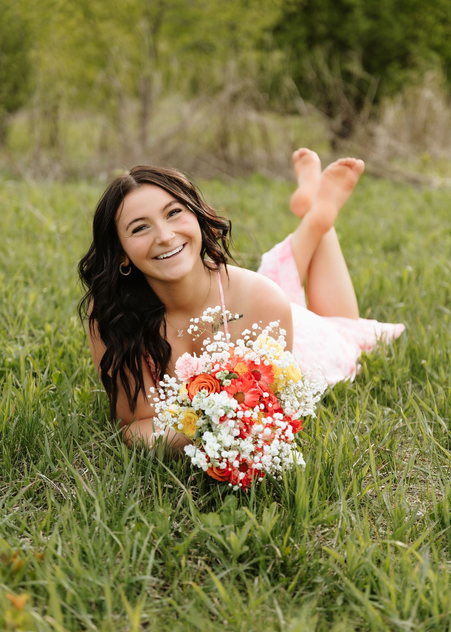 Young woman in pink dress lying in green grass, smiling, holding colorful bouquet of flowers outdoors.