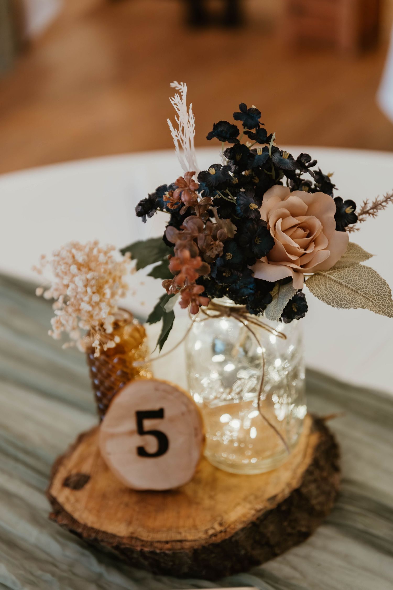 Rustic wedding centerpiece with table number 5 on wood slice featuring blush roses and black flowers in mason jar.