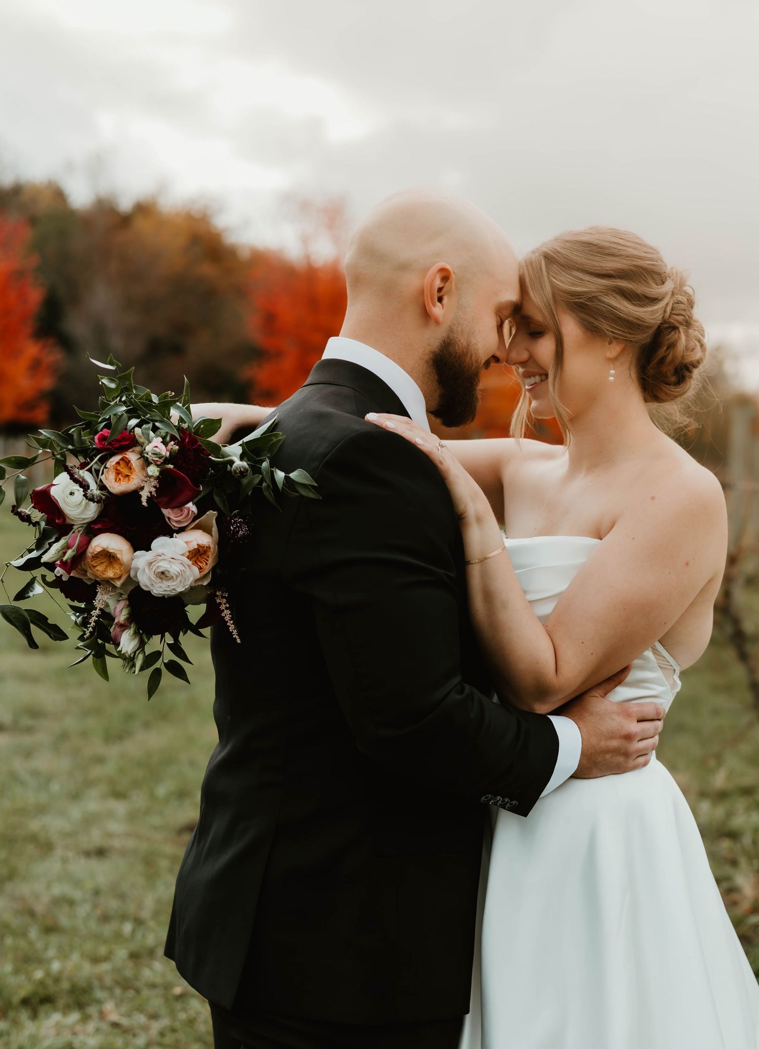 Newlyweds share an intimate moment against autumn foliage while holding a floral bouquet.