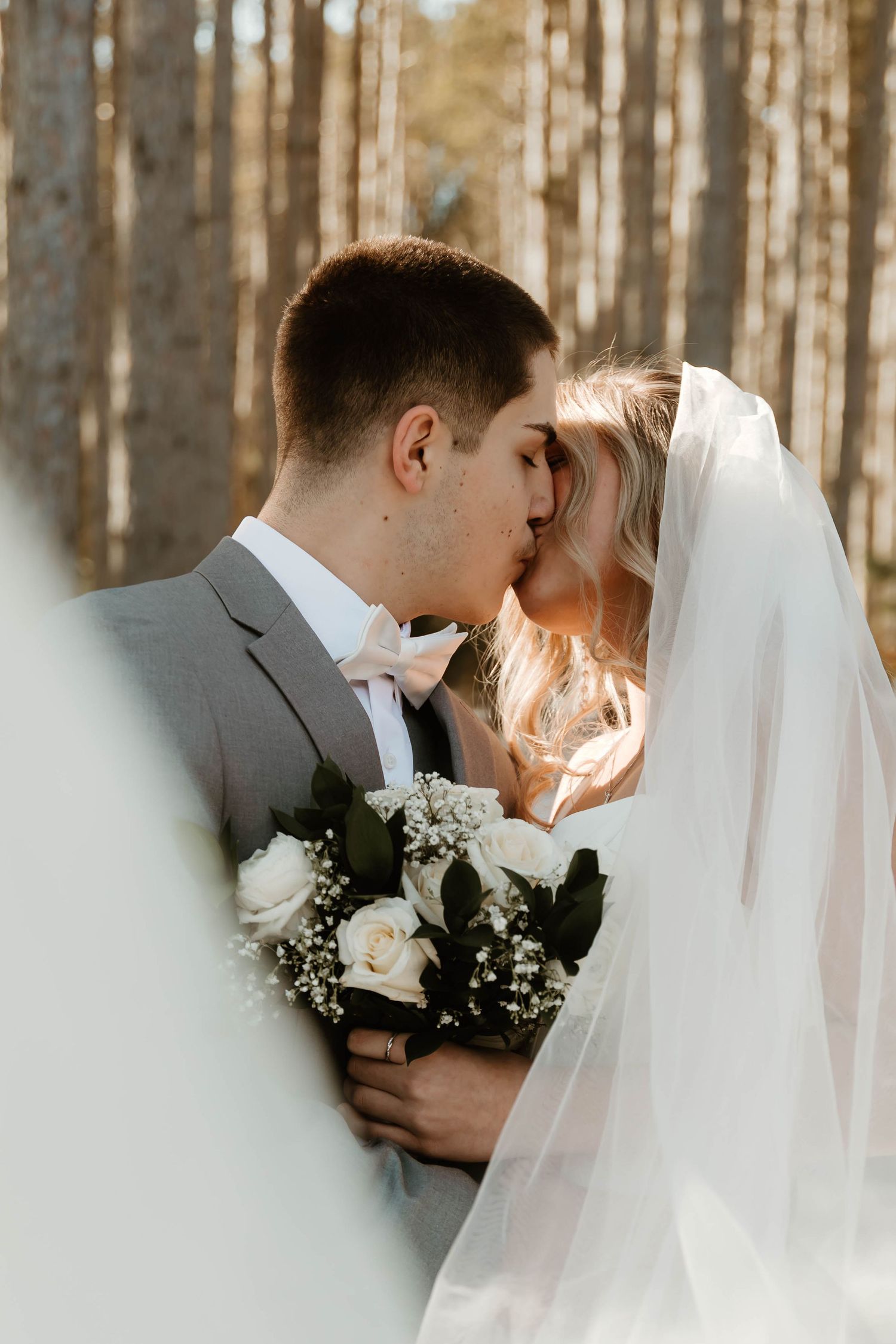 A romantic winter wedding moment captured in a forest with sunlight filtering through the trees.