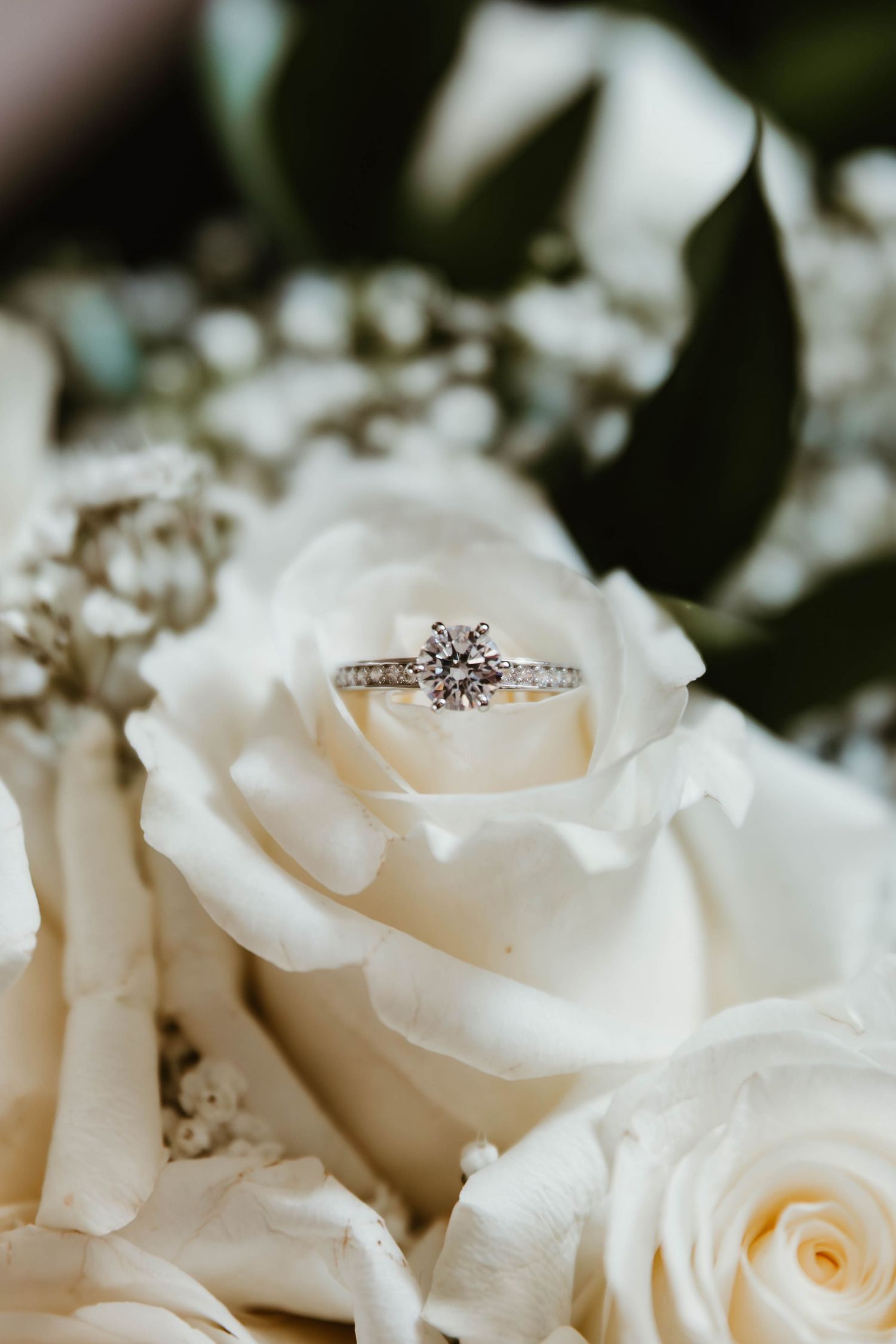 A sparkling diamond engagement ring nestled in the center of a creamy white rose bloom among a bridal bouquet.