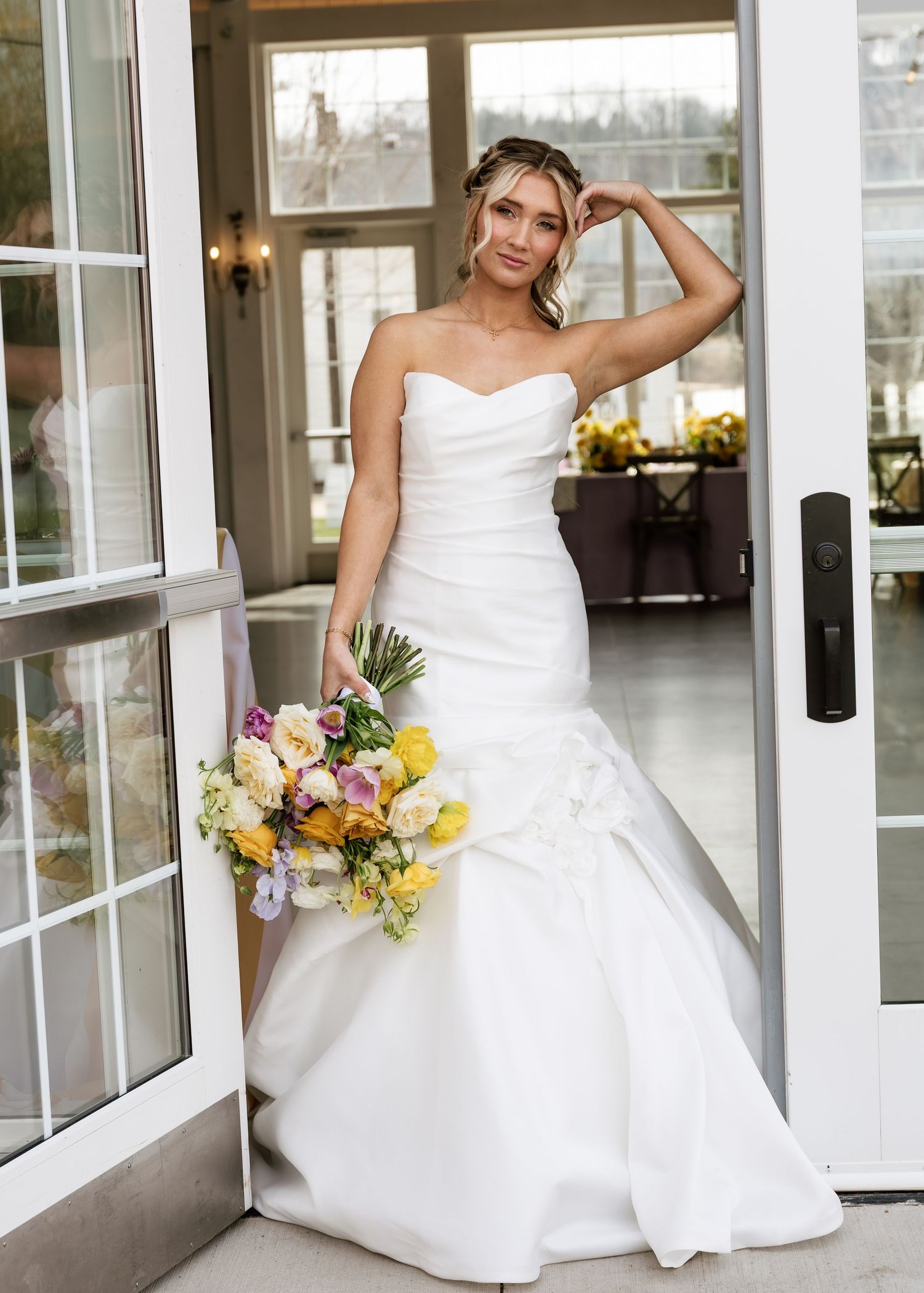Bride in strapless white mermaid wedding gown holding colorful bouquet, posing in doorway of elegant venue.