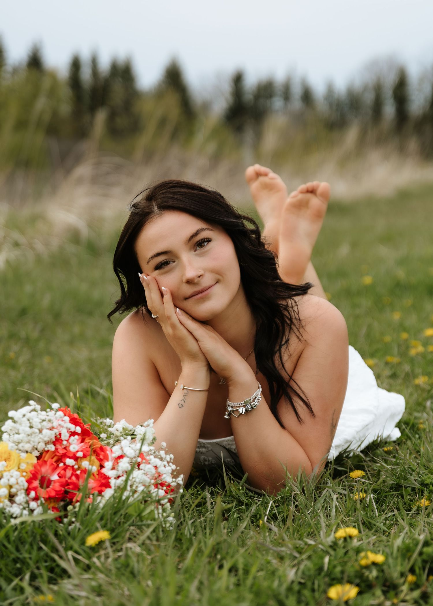 Young woman in white dress lying on grass with colorful flower bouquet, smiling peacefully outdoors.