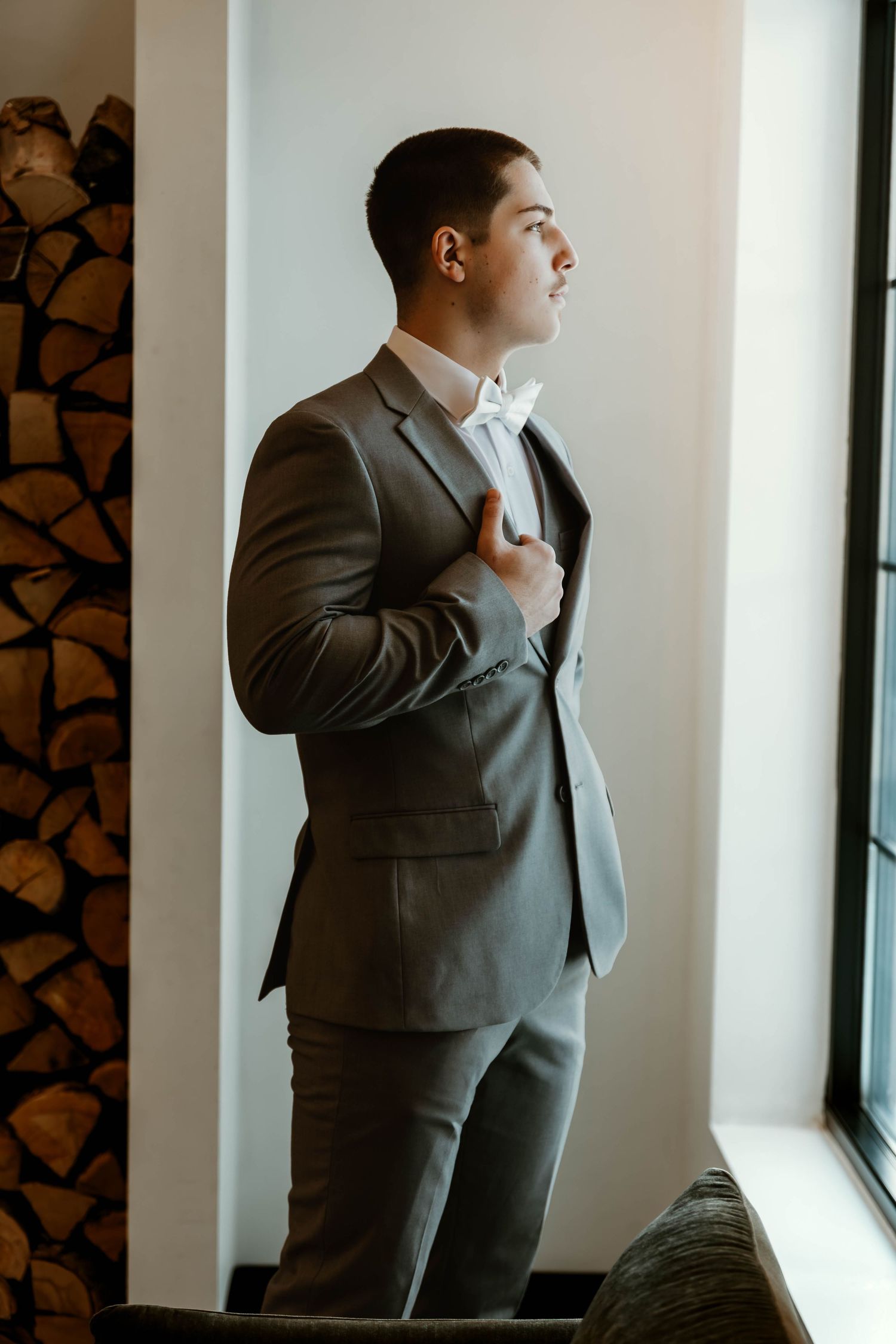 Figure in a grey suit adjusting tie while standing by a window.