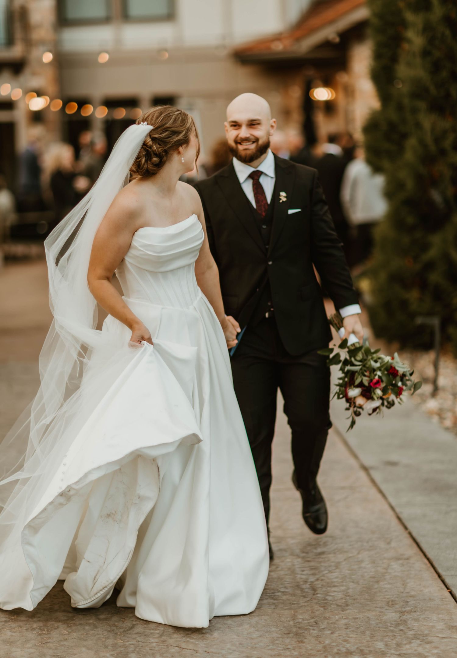 Bride's dress flows gracefully as couple walks along pathway with string lights overhead.