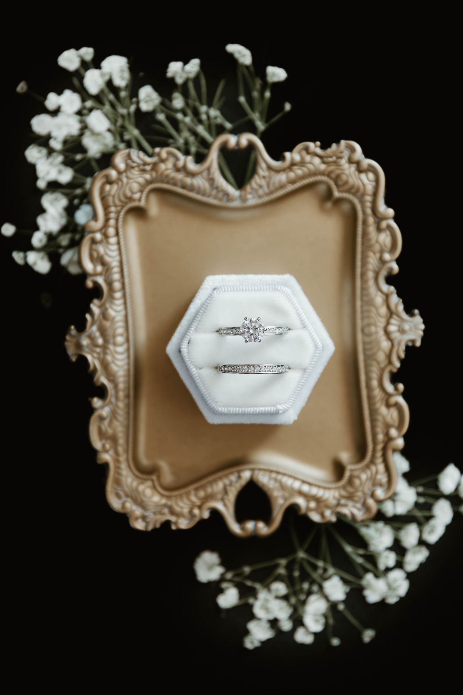 Wedding rings displayed in hexagonal white boxes on ornate gold vintage trays with baby's breath accents.