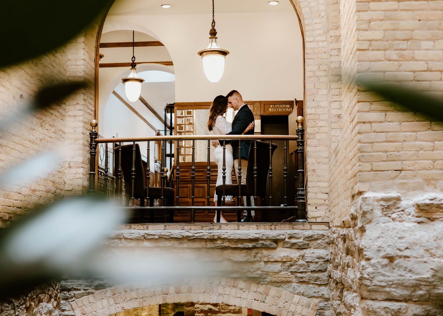 A romantic couple shares a tender moment in a rustic brick-walled venue with vintage lighting and wooden beams.