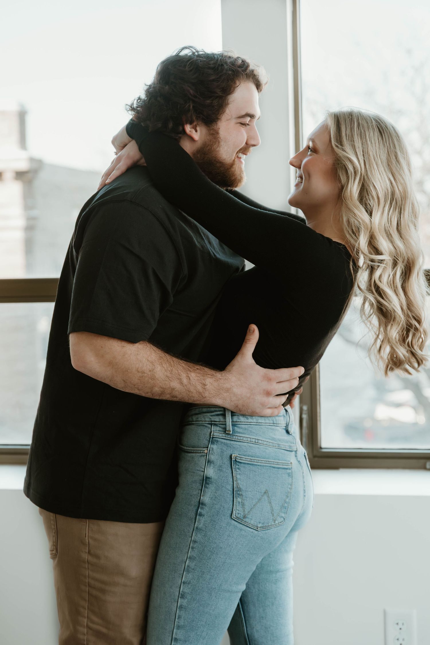 Romantic couple embracing by window in casual attire showing affection and intimacy.