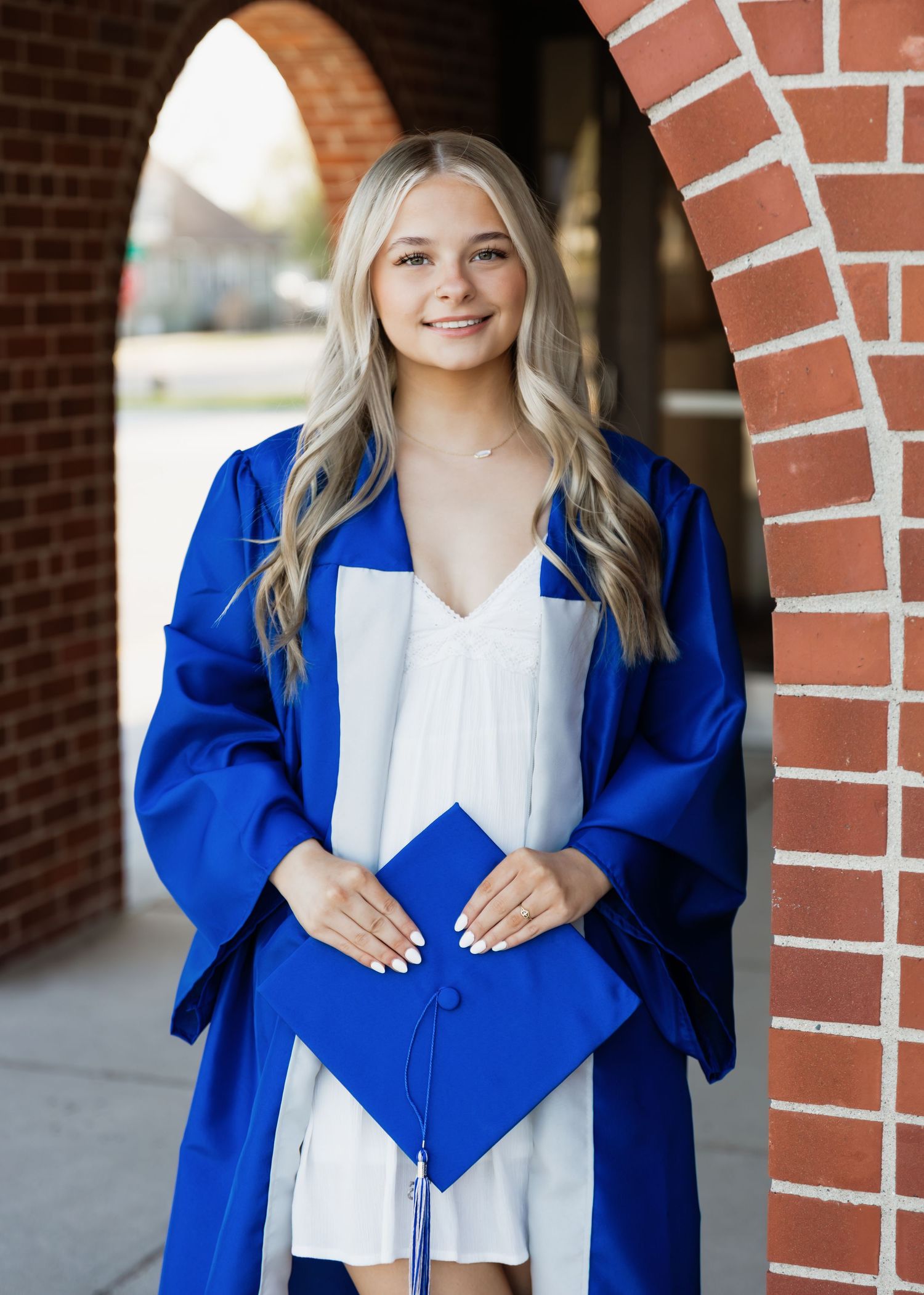 Young woman in blue graduation cap and gown smiling outdoors near brick archway.