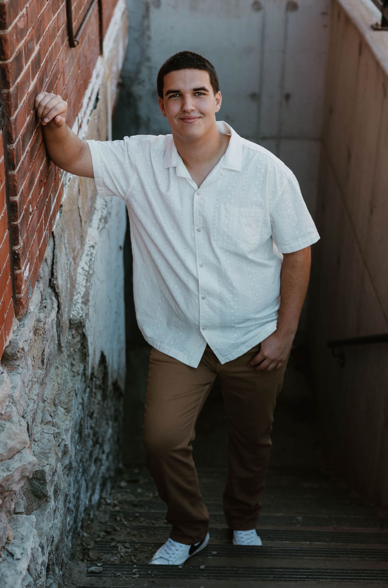 Young man in white shirt and brown pants leaning against brick wall on outdoor steps.