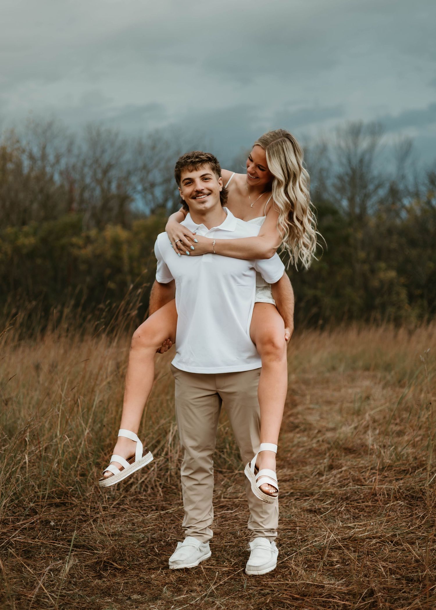 A playful piggyback ride moment captured in a golden field with moody overcast skies.