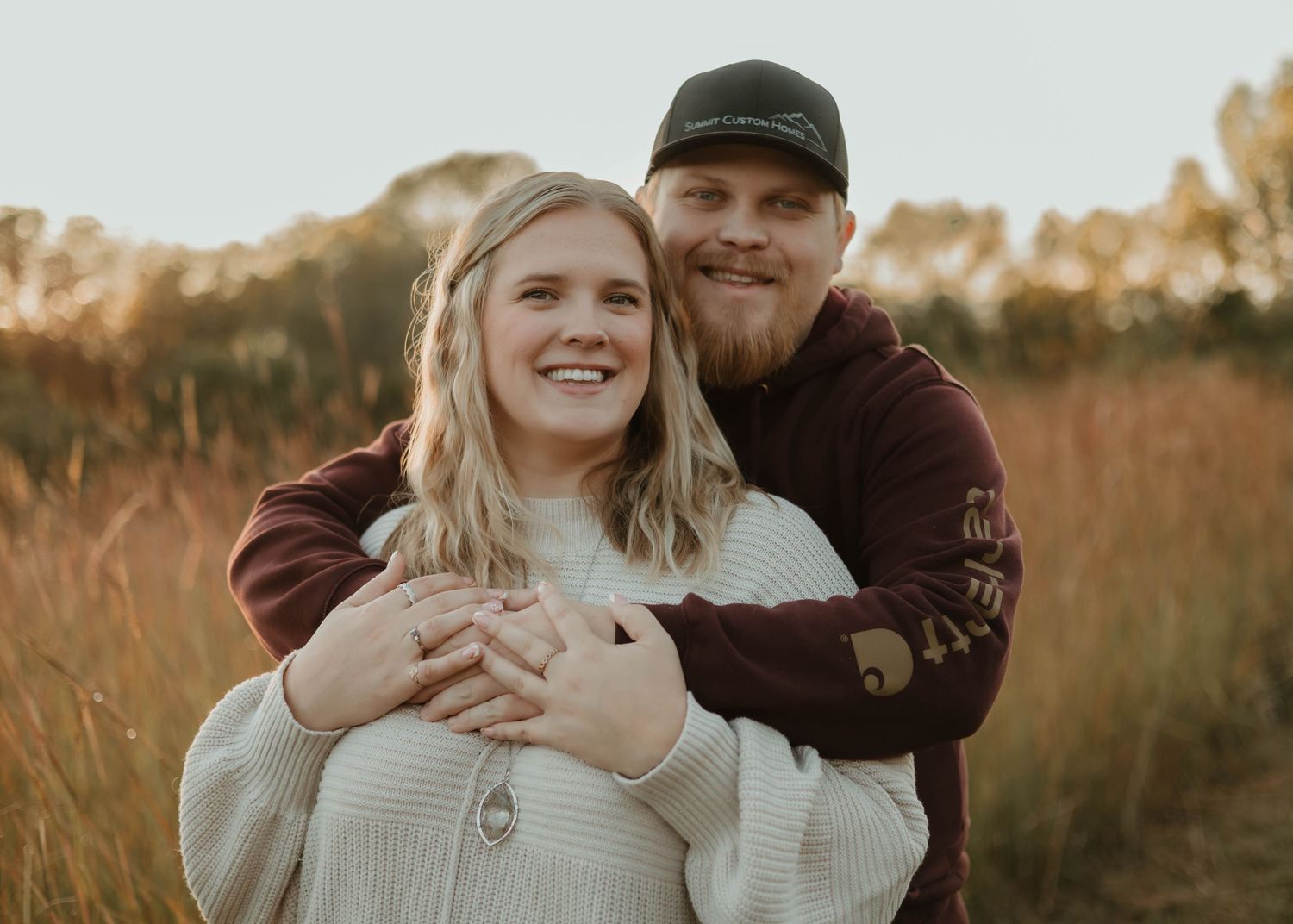 Couple embraces in a golden field during sunset wearing cozy sweaters and casual attire.