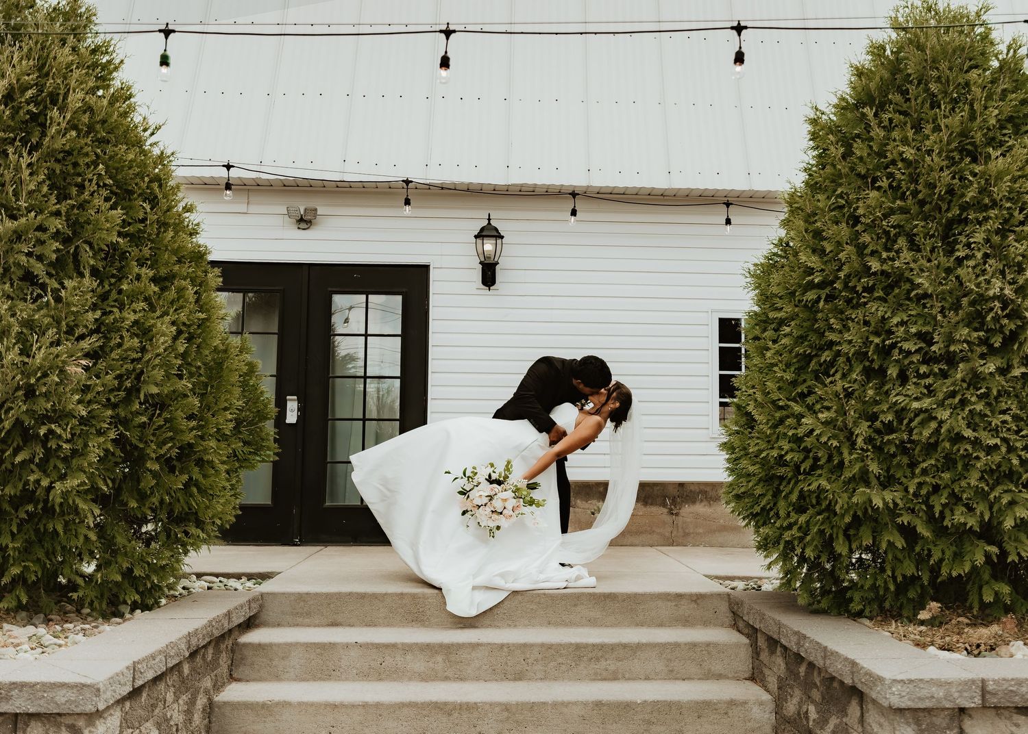 Bride and groom sharing a romantic dip kiss on stone steps outside a white farmhouse venue with lush green trees.