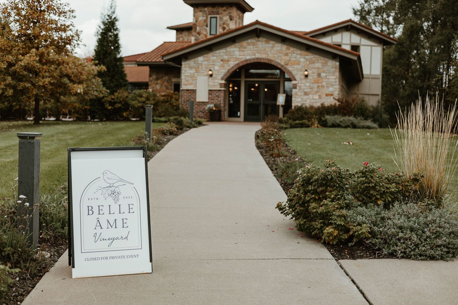 A curved concrete path leads to a rustic stone and wood building with a Belle Ame sign in the foreground.