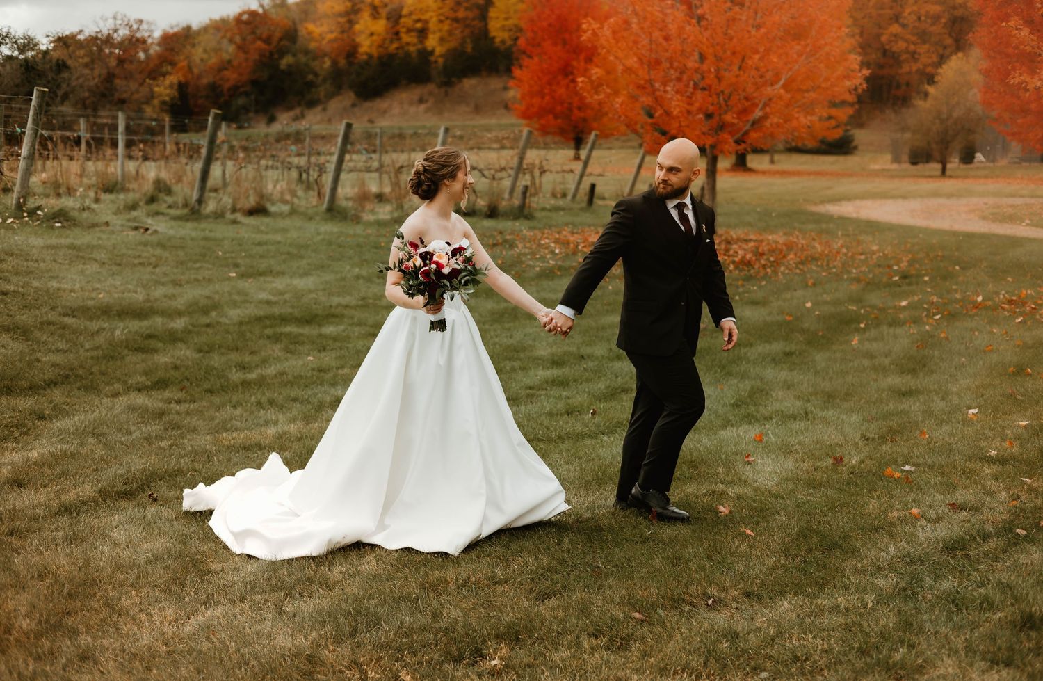 A couple in wedding attire walks through a scenic autumn landscape with vibrant orange foliage in the background.