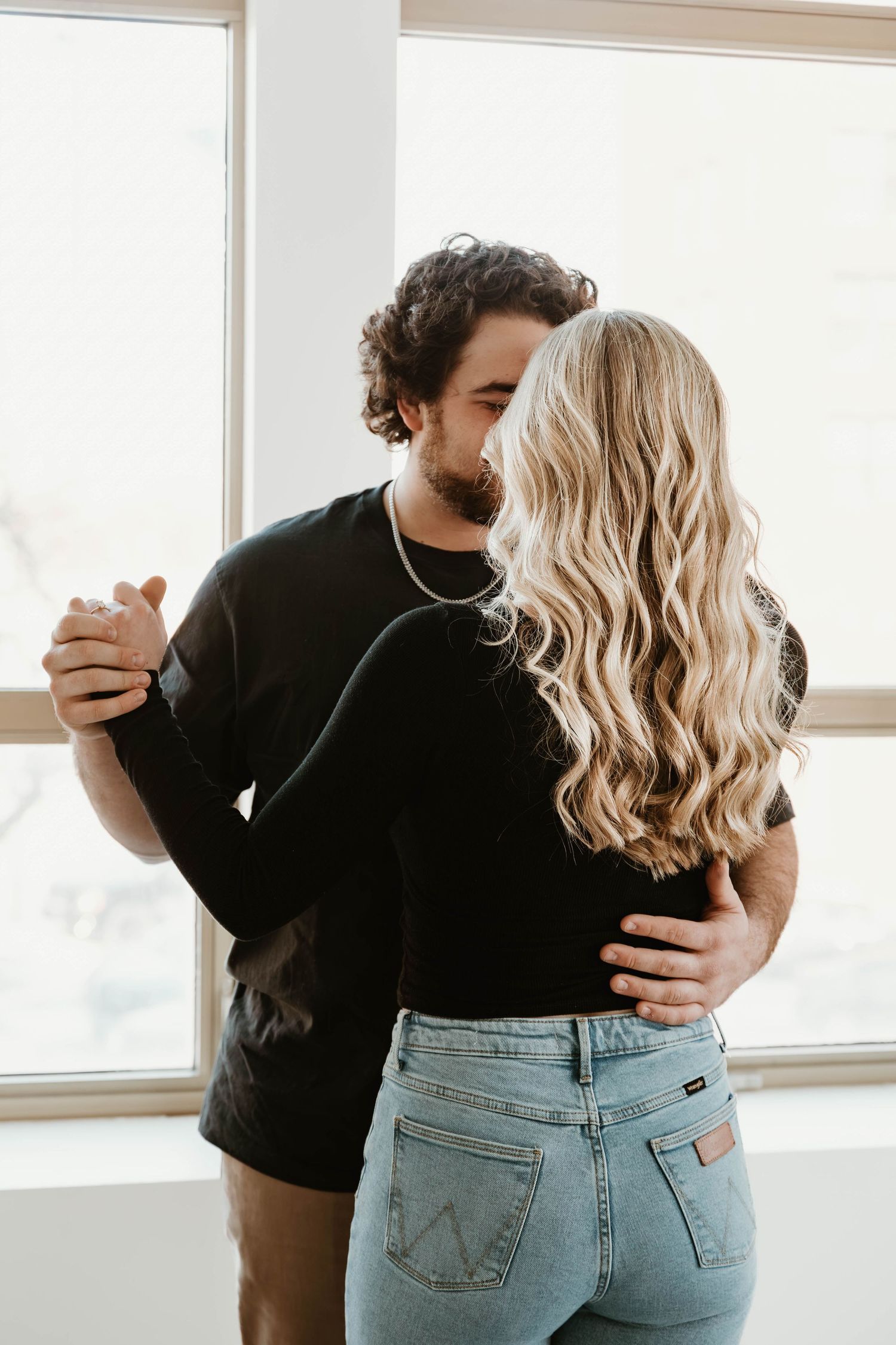 Tender moments between a couple in black and denim are captured near a sunlit window.
