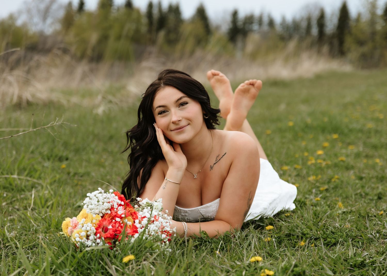 Young woman in white dress lying on green grass, smiling, holding colorful wildflower bouquet outdoors.