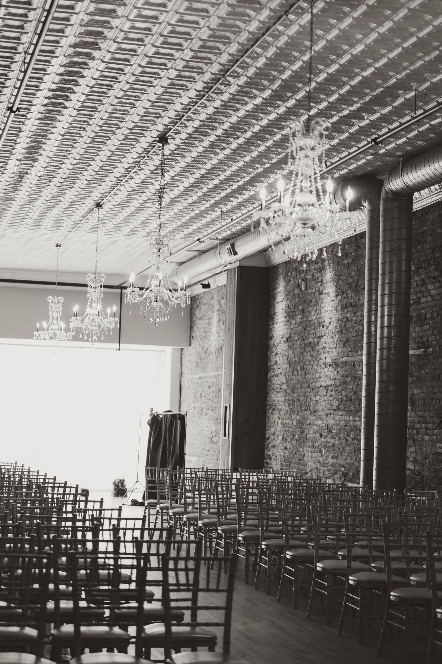 Elegant wedding ceremony space with rows of dark chairs, crystal chandeliers, and exposed brick walls.