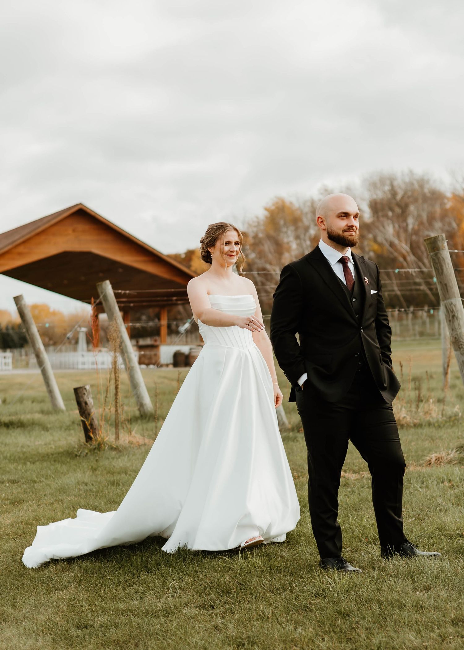 Wedding couple posing in rustic outdoor setting with wooden structures and autumn scenery.