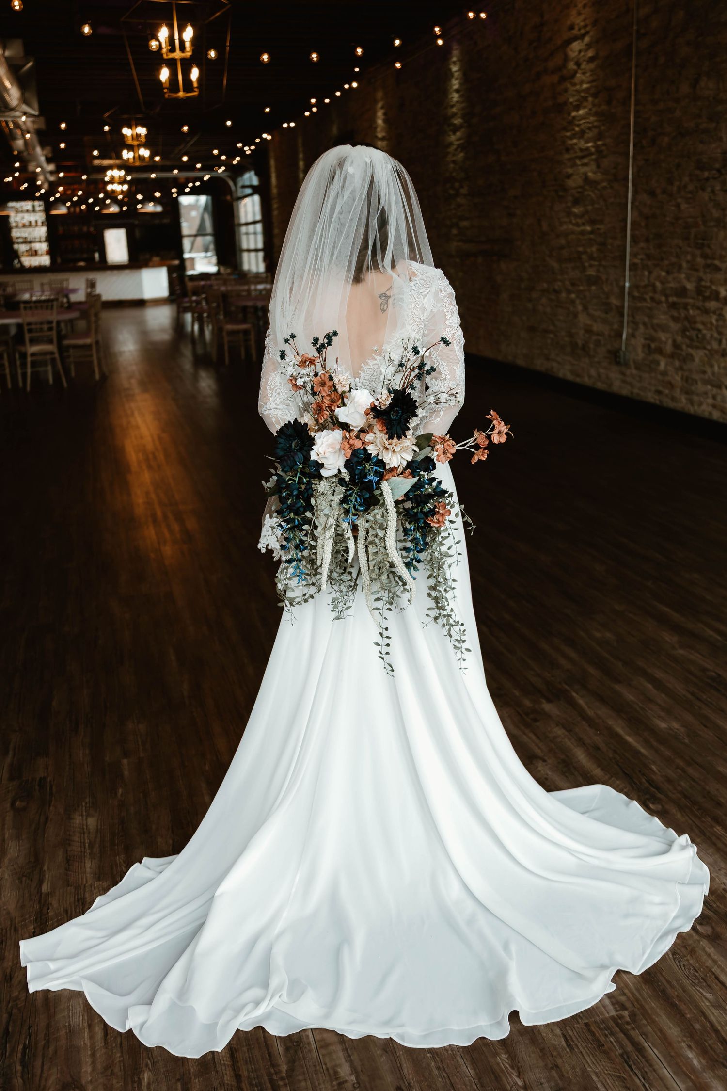 Bride in white dress holding bouquet against dark rustic venue with string lights overhead.