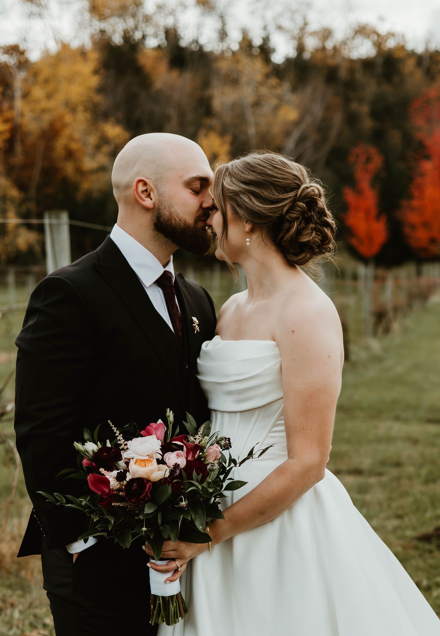 A couple embraces while holding a bouquet of dark flowers in an autumn vineyard.