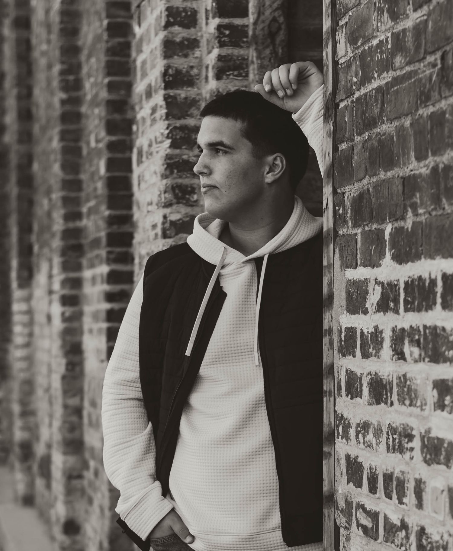 Young man in white hoodie and black jacket leaning against a brick wall in a black and white photo.