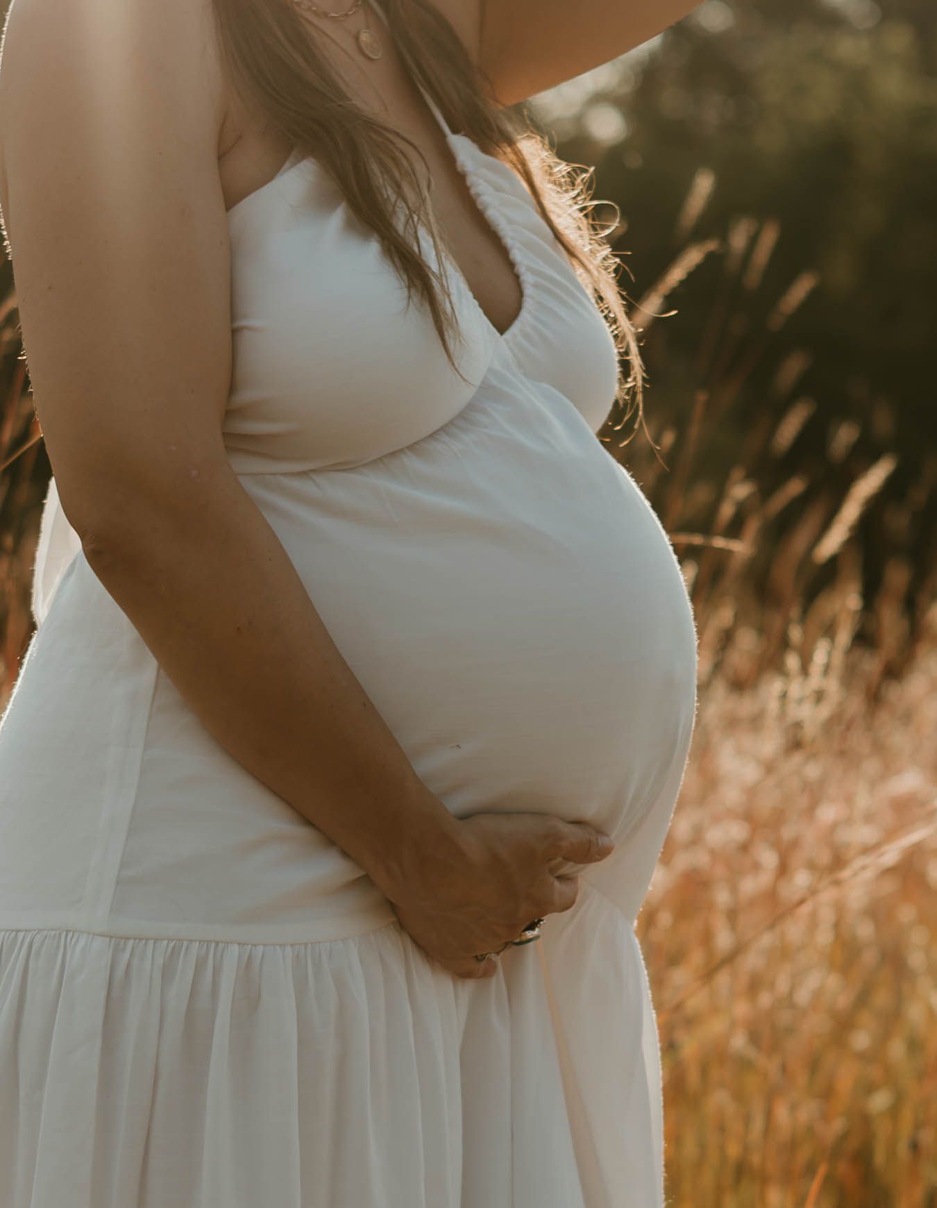 Side view of pregnant woman in white dress standing in sunlit field of tall grass.