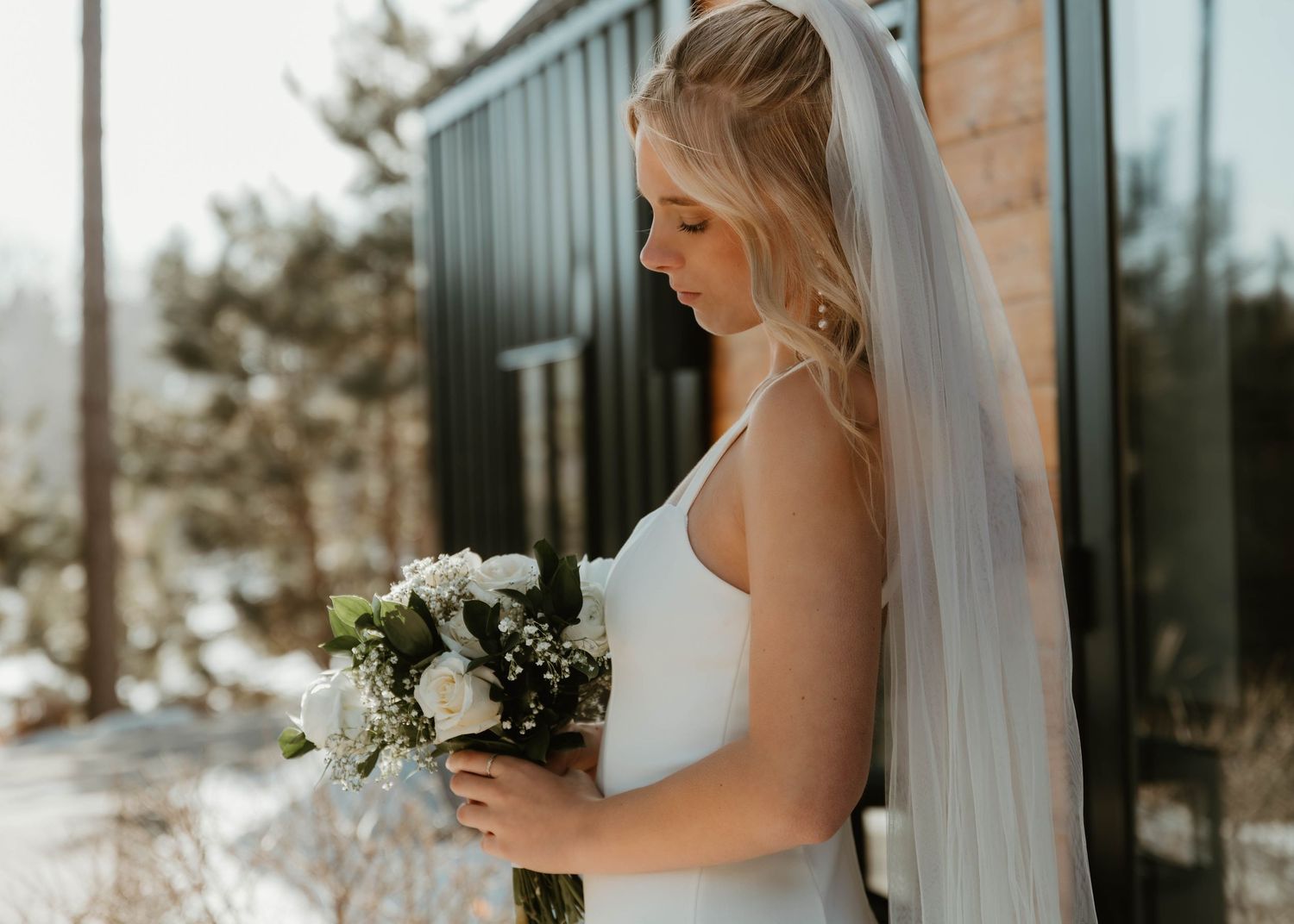 Bride in white wedding dress holding bouquet stands against rustic brick wall in natural sunlight.